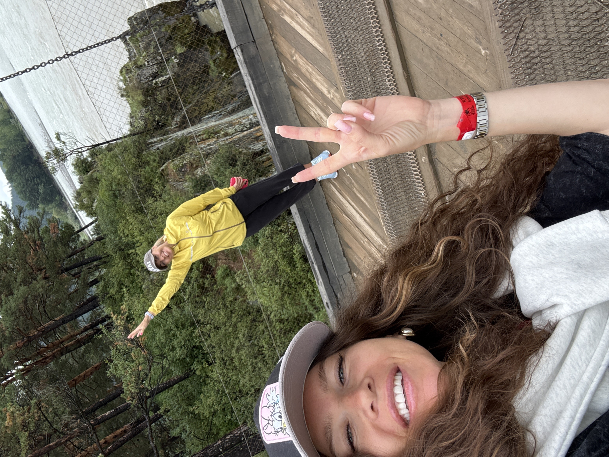 The image depicts two women on a wooden walkway overlooking a river. The woman in the foreground is smiling and looking at the camera, wearing a black baseball cap and a white scarf. Her right arm is raised, making a peace sign with her fingers. She has long, curly brown hair and is wearing a watch and a red wristband on her left wrist. The second woman, in the background, is wearing a yellow jacket and black pants, with a grey baseball cap. She is smiling and raising her left arm. The environment is characterized by lush green trees and a rocky cliff face near a wide, flowing river. The walkway appears to be part of a bridge or elevated pathway.