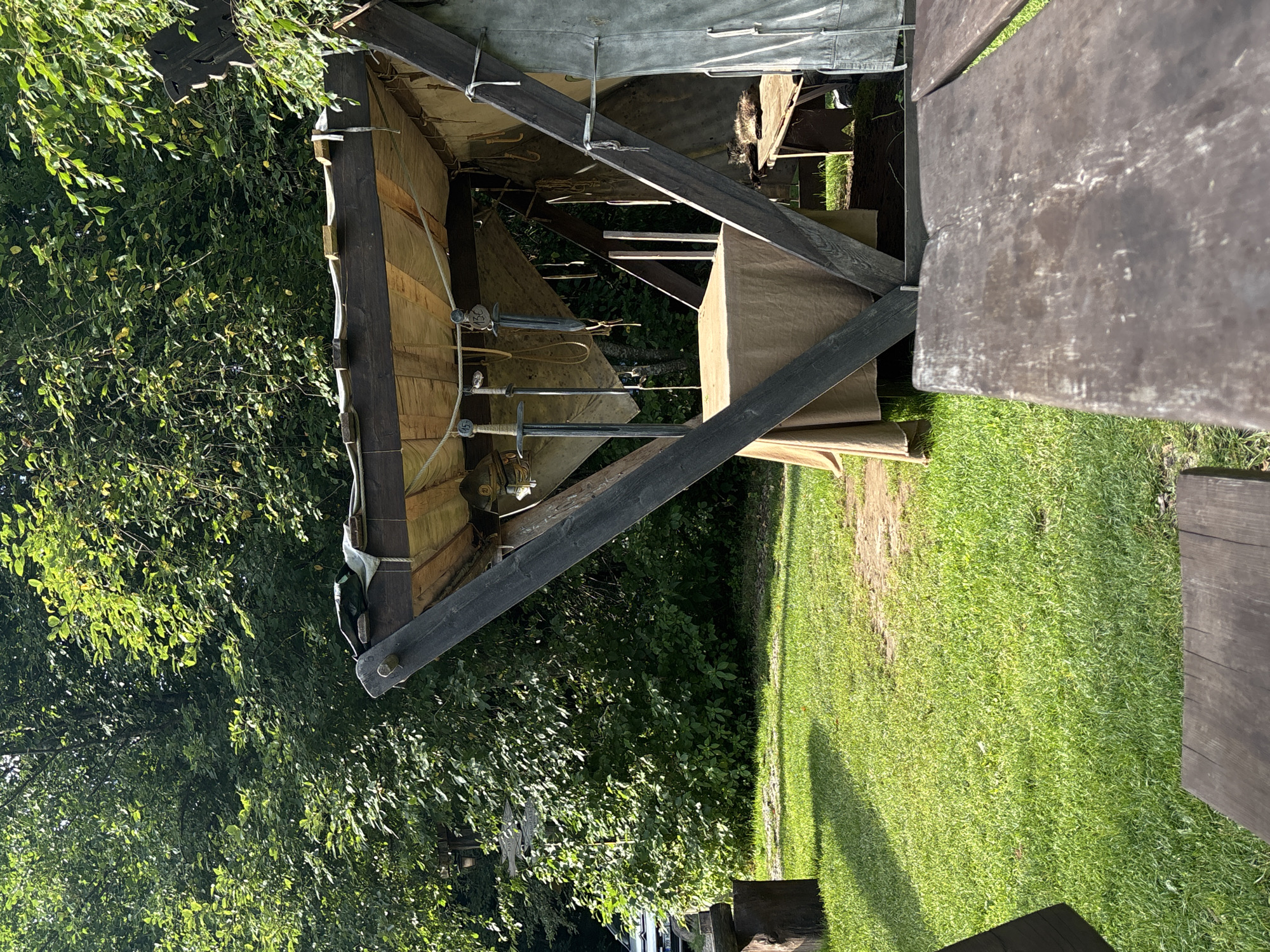 In Saula, Estonia, a scene unfolds with a rustic, open-air market stall set against a backdrop of lush green trees. The stall, constructed from dark wooden beams and a tan canvas roof, displays several medieval-style swords hanging from the ceiling. A table draped in a tan cloth occupies the center of the stall. To the right, a bench is occupied by a tabby cat lounging peacefully. The ground around the stall is covered in neatly trimmed green grass, with a weathered wooden table in the foreground, suggesting a leisurely outdoor setting.