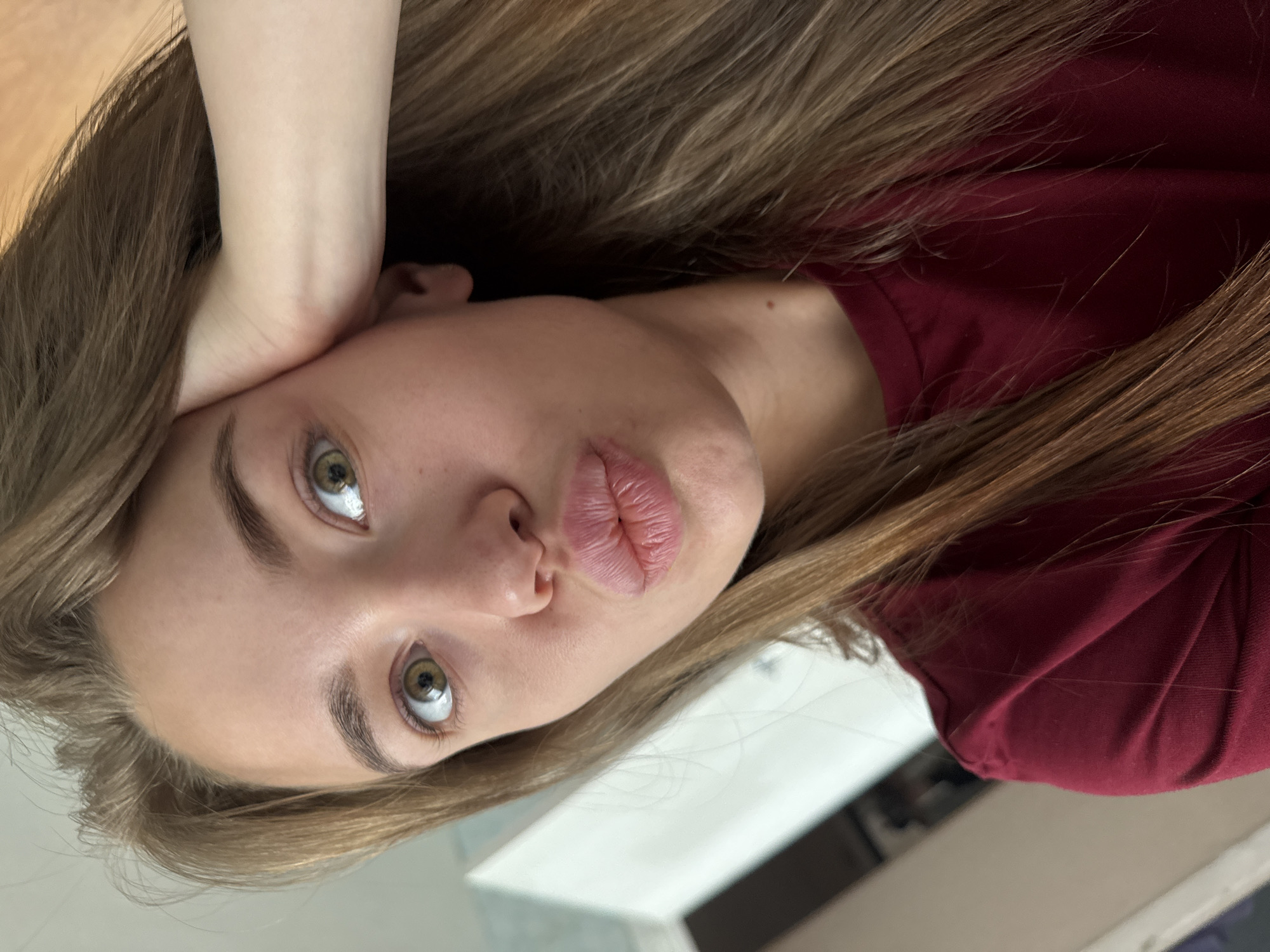 A young woman with long brown hair and green eyes is taking a selfie indoors. She is wearing a maroon t-shirt and has her right hand on her head. Her lips are pursed. The background is out of focus but appears to be a room with a white wall and door.