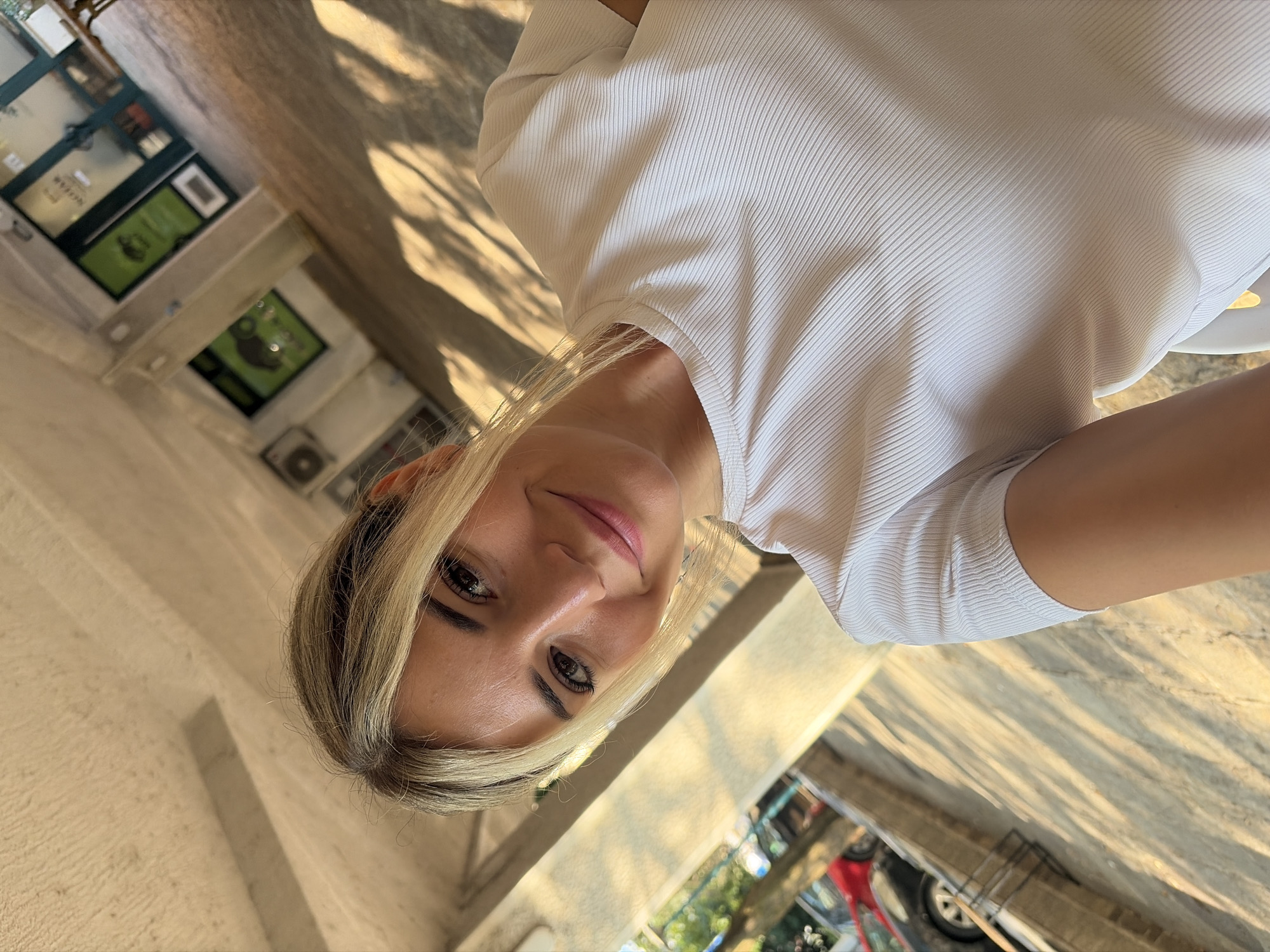 A woman with blonde hair is captured in a selfie taken outdoors, likely in Budva, Montenegro, as indicated by the visual cues of a Mediterranean setting. She is smiling and looking directly at the camera, her face illuminated by natural light. She is wearing a white, ribbed short-sleeved shirt. The background shows a building with windows and doors, a walkway with patterned shadows from sunlight, and some greenery and a red car visible on the left. The architectural style suggests it could be part of a commercial area or an older building in a coastal town.
