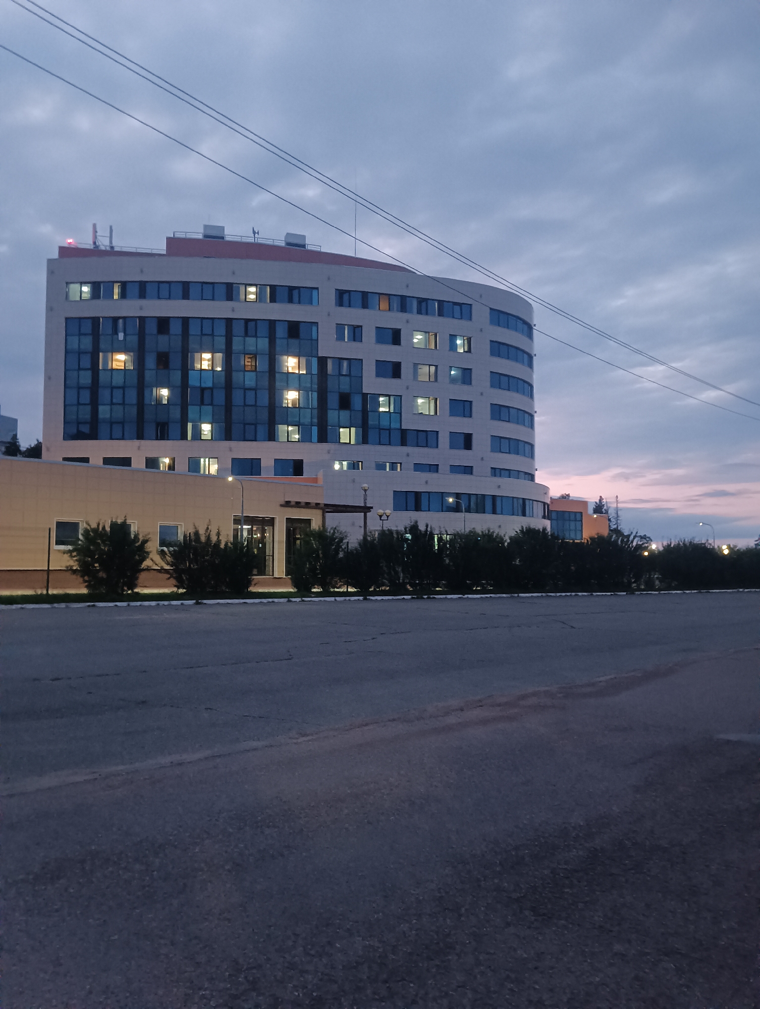 A multi-story office building with a curved facade is pictured at dusk on a cloudy day. The building, located in the Privolzhsky district of Perm Krai, Russia, features large windows that are illuminated from within, suggesting interior lighting is on. A paved road is in the foreground, with a row of bushes and trees running along the base of the building. Power lines are visible in the sky. The overall scene depicts an urban environment transitioning from day to night.