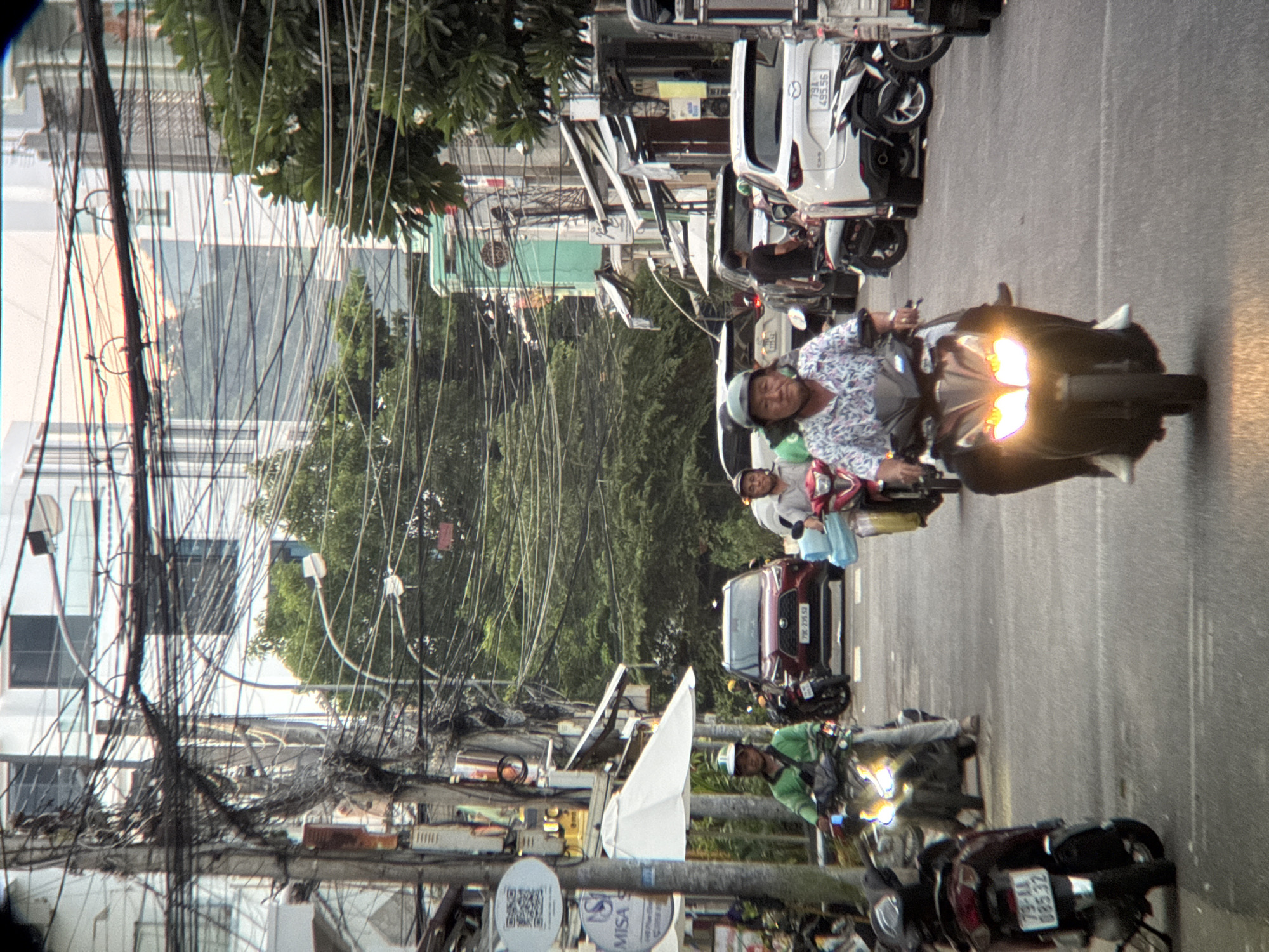 This image depicts a street scene in Nha Trang, Vietnam, characterized by a dense network of overhead electrical wires. In the foreground, several individuals are riding motorcycles. A person in a patterned shirt and dark pants rides a dark-colored motorcycle with illuminated headlights, moving towards the viewer. To the left, a person wearing a green long-sleeved shirt and a helmet rides a motorcycle. Further in the background, a red SUV with license plate "79C-225.52" is visible, along with other vehicles and motorcycles. Commercial buildings with awnings line the street on the right. Lush green trees are present on both sides of the street, contrasting with the urban infrastructure. The overall impression is of a busy urban environment with typical street life in Vietnam.