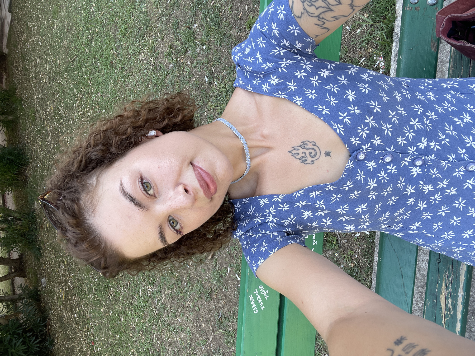 A young woman with curly brown hair and green eyes is sitting on a green park bench and taking a selfie. She is wearing a blue dress with a white floral pattern and a light blue choker necklace. She has a tattoo on her chest of flames and another tattoo on her arm. The bench has some graffiti written on it in white. She is sitting in front of a grassy area with trees in the background. The scene appears to be in a park in Budva, Montenegro.