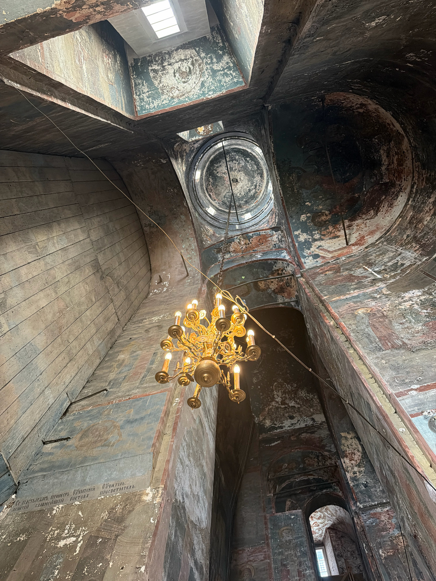 The image shows the interior of an old church in Novo-Nikolskoye, Russia. The camera is looking upwards, capturing the ceiling, dome, and the upper parts of the walls. A large, ornate brass chandelier with multiple lit bulbs hangs prominently in the center of the frame. The walls and ceiling are decorated with faded, peeling frescoes, depicting religious figures and scenes. The architecture is complex, with arched openings and a multi-levelled ceiling structure. The overall impression is one of age and historical significance, with a somber yet majestic atmosphere. Natural light enters from a skylight at the top. The wooden planks on the left wall are visible, showing their aged texture and horizontal arrangement. The frescoes on the walls are partially obscured by wear and tear, but their outlines and colors are still discernible. The scene is static, with no people or active events taking place. The environment appears to be a preserved historical site.