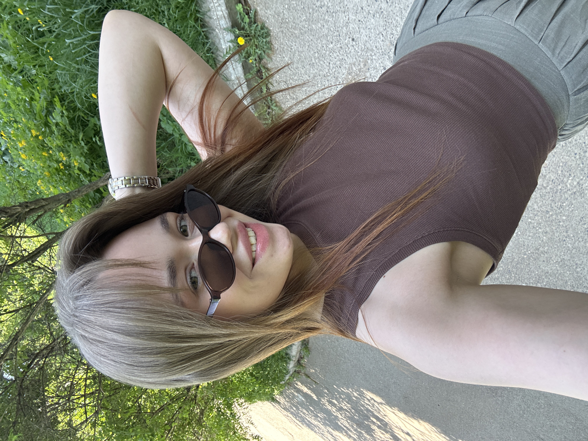 A young woman with long, light brown hair, wearing sunglasses, a brown tank top, and a pleated grey skirt, poses for a selfie outdoors. She is on a paved surface next to a grassy area with trees and yellow flowers. The exact location within the city of Chaikovsky, Russia is not discernible.