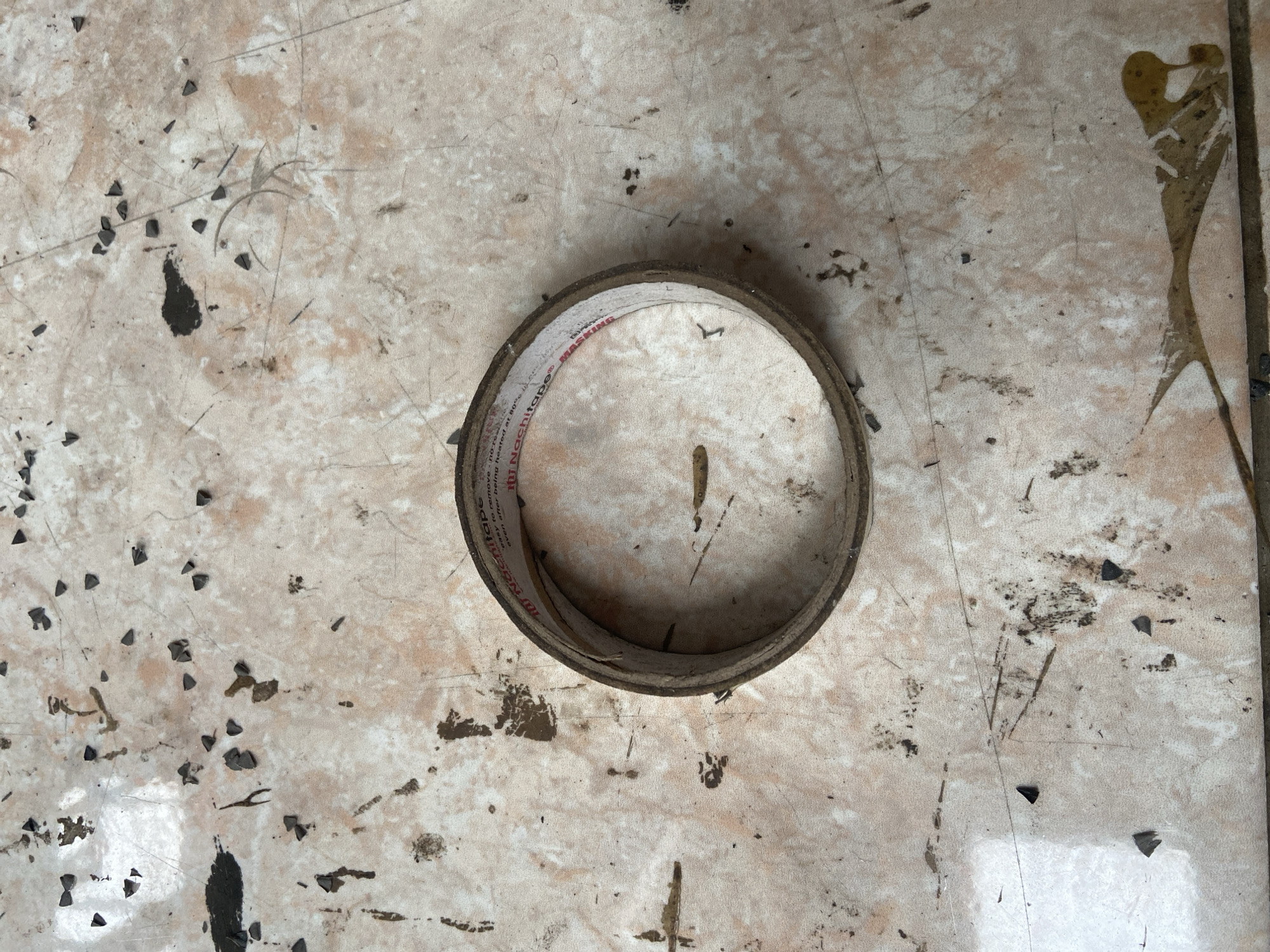 A roll of masking tape is lying on a tiled floor with various debris scattered around. The tape roll is circular and made of paper, with red lettering visible on its surface. The tiles are a light color with reddish-brown streaks, and there are numerous small, dark, triangular pieces of debris, along with some larger dark smudges and a yellow liquid stain. The overall scene appears to be outdoors, possibly on a patio or walkway, given the presence of debris and the outdoor lighting. The image was captured at a street address in Plosokandang, Indonesia.