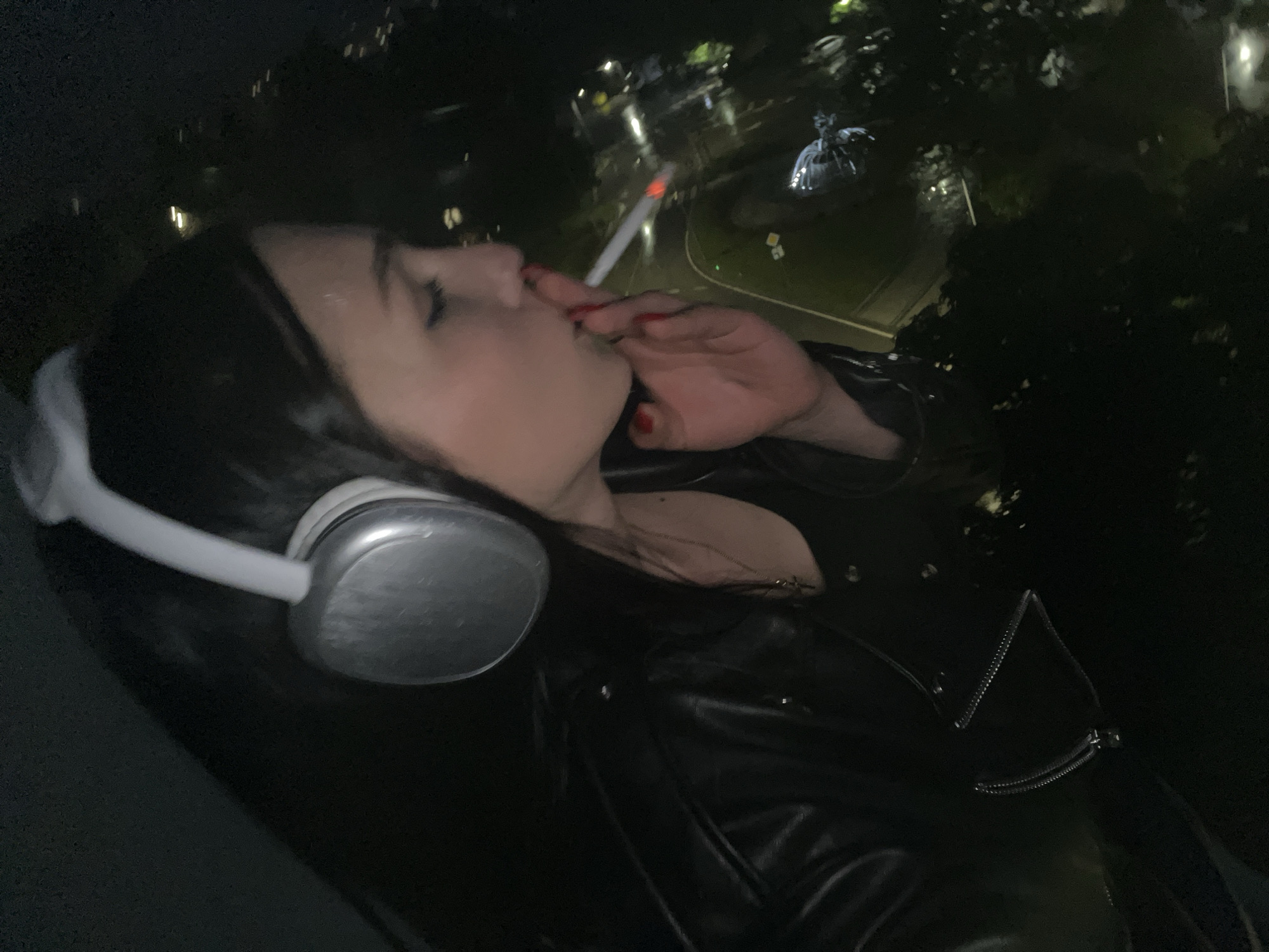 A dimly lit nighttime image captures a person, likely female, from a high vantage point, smoking a cigarette and wearing headphones. The subject is seen in profile, angled downwards, with dark hair and a light complexion. She is wearing large, silver or gray over-ear headphones, and a dark, possibly leather, jacket with a visible zipper. Her fingernails are painted red, and she holds a lit cigarette to her mouth. A delicate necklace with a small pendant is visible around her neck.
The background is dark, indicating night, and features numerous scattered lights that suggest an urban or suburban environment. Below and to the right of the subject, illuminated trees and a distinct, light-colored object or structure, possibly a statue or sculpture, are visible, further suggesting an outdoor public space like a park viewed from above. The overall scene conveys a quiet, contemplative moment in a city at night. No visible text is present in the image.
