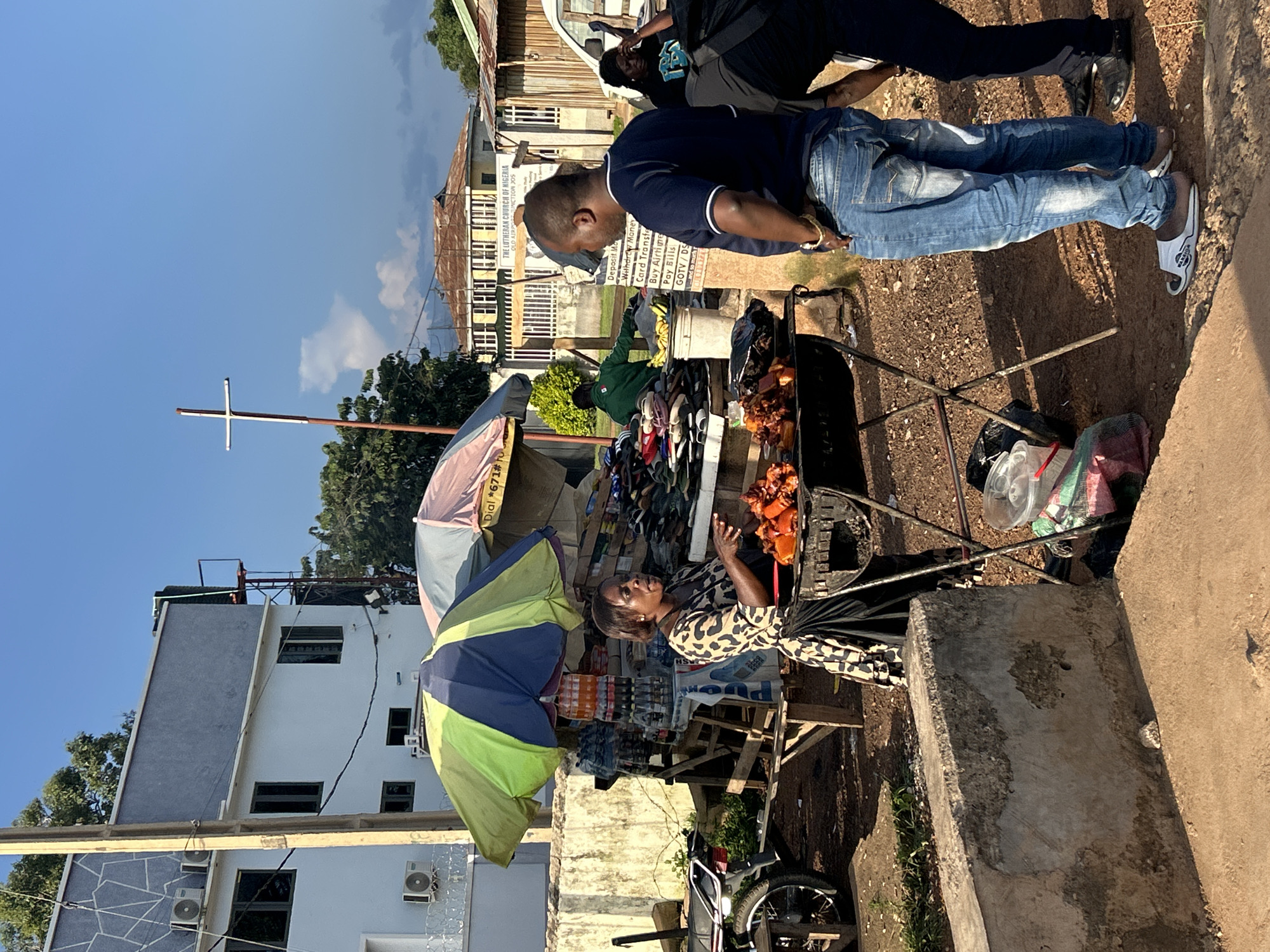 In the city of Jos, Nigeria, an outdoor market scene unfolds under a clear blue sky. A woman, dressed in a leopard-print dress and a black skirt, stands behind a grill, attending to a pile of cooked meat. In front of her, a man in a dark blue t-shirt and light blue jeans observes the food, his left arm resting on his hip. To his right, another person, partially visible, wears a black shirt and pants. The market stall displays various items, including drinks in bottles, and an assortment of shoes. A green and yellow umbrella partially shades the stall. In the background, buildings of varying architectural styles are visible, including one with a prominent cross on a pole. A white van is parked to the right. The ground is composed of dirt and gravel.