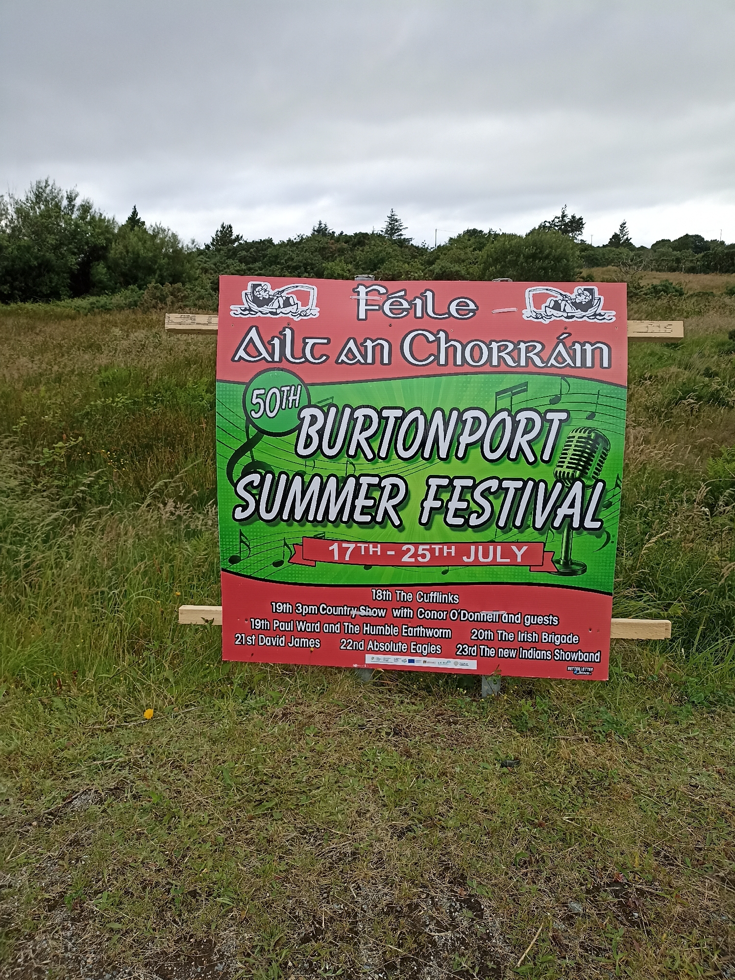 A large, red and green poster advertising the "BURTONPORT SUMMER FESTIVAL" is mounted on wooden planks outdoors. The poster features a vintage microphone graphic, musical notes, and text in English and Irish. The sky is overcast, and the poster is situated in a grassy area with trees in the background. The bottom of the poster lists various events and performers, including "19th 3pm Country Show with Conor O'Donnell and guests," "19th Paul Ward and The Humble Earthworm," "20th The Irish Brigade," "21st David James," and "22nd Absolute Eagles," and "23rd The new Indians Showband." The festival is scheduled to take place from the 17th to the 25th of July.
