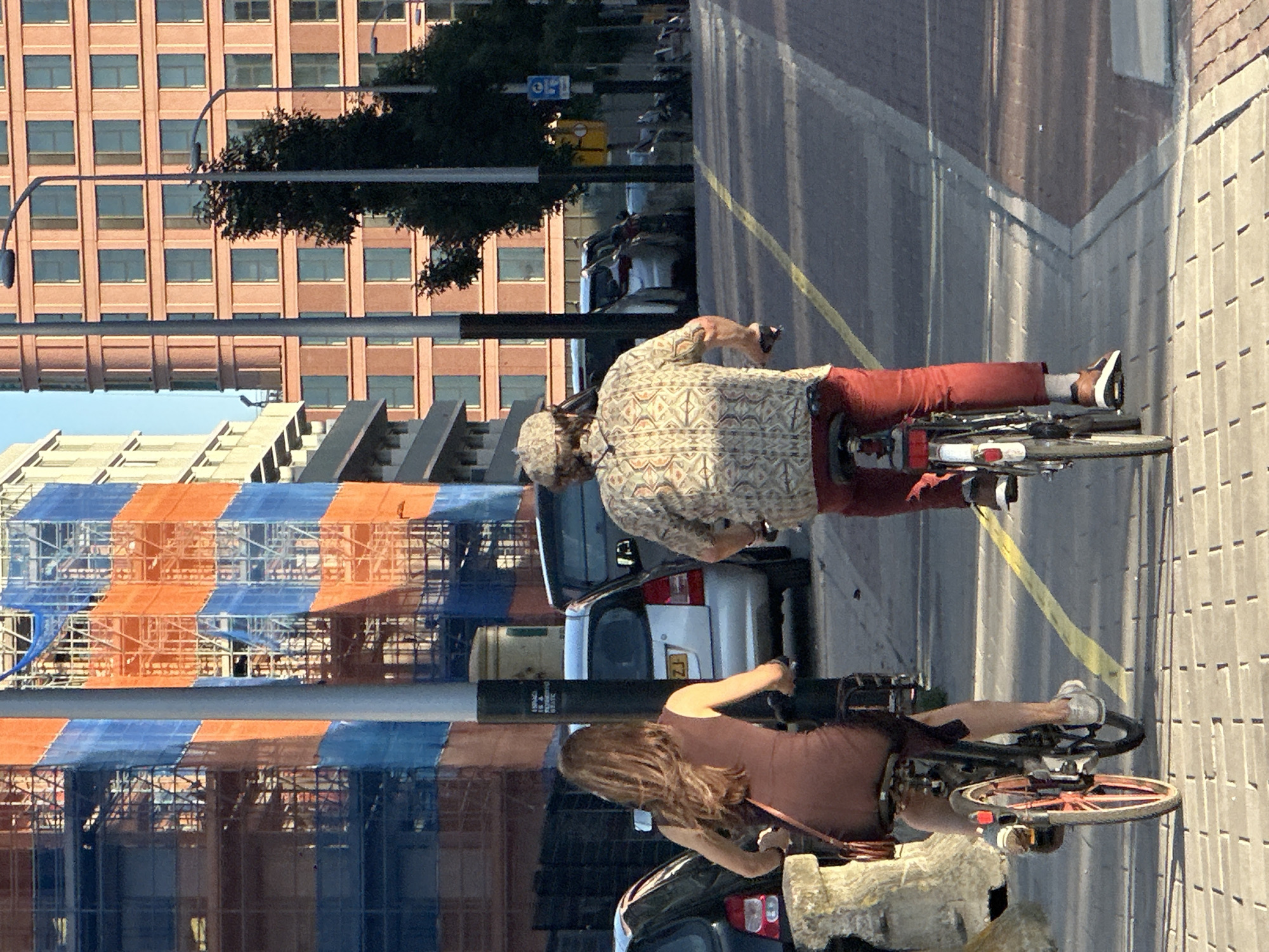 This image shows two people on bicycles in what appears to be a city. The scene is a street with buildings and construction scaffolding visible. The time of day appears to be daytime, likely afternoon, given the shadows cast. The weather seems to be clear and sunny.
In the foreground, a woman with long brown hair is riding a bicycle. She is wearing a brown tank top and appears to be holding onto the handlebars. Her bicycle has a red rim on the wheel.
Further back, a person wearing a patterned shirt and red pants is riding another bicycle. This person is also holding onto the handlebars. The bicycle appears to have a rear rack with a red cover.
Behind the cyclists, there are buildings with numerous windows and construction scaffolding covered with orange and blue netting. Cars and other vehicles are also visible in the background. A yellow sign with blue lettering is partially visible.
The image is oriented vertically, suggesting it might have been taken with a phone camera or that the original orientation was rotated. The overall impression is a snapshot of everyday life in an urban environment, possibly a tourist or local cycling through the city.