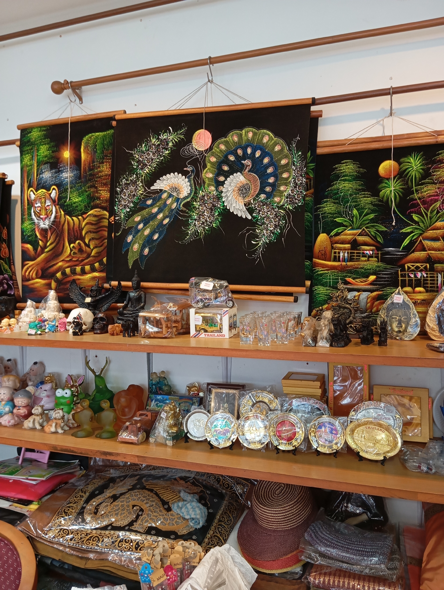 The image shows a souvenir shop in Karon, Thailand. The shop is filled with various items, including tapestries, statues, glassware, and decorative plates.
In the upper part of the image, three large tapestries are hanging from a wooden rod. The leftmost tapestry depicts a tiger. The middle tapestry features two peacocks, and the rightmost tapestry shows a tropical village scene.
Below the tapestries, on a wooden shelf, are various decorative items. These include small statues of elephants, Buddha heads, and what appear to be mythological creatures. There are also several shot glasses and small boxes.
The lower shelf is filled with a variety of items. These include stuffed animals, ceramic figurines, a large decorative tapestry featuring a dragon, and several woven hats. There are also stacks of packaged items.
The overall impression is of a well-stocked souvenir shop offering a range of items typical of Thai tourism.
