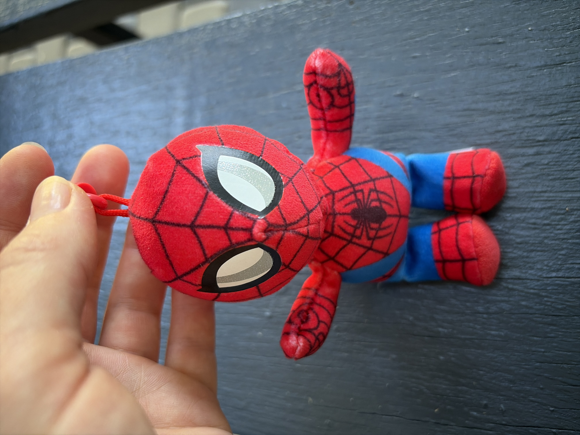 A person is holding a plush Spider-Man toy by a red loop. The toy is red and blue with black spiderweb markings. The background is a dark, textured surface, likely a table. The scene does not provide information to determine its location in Oxentea, Moldova.