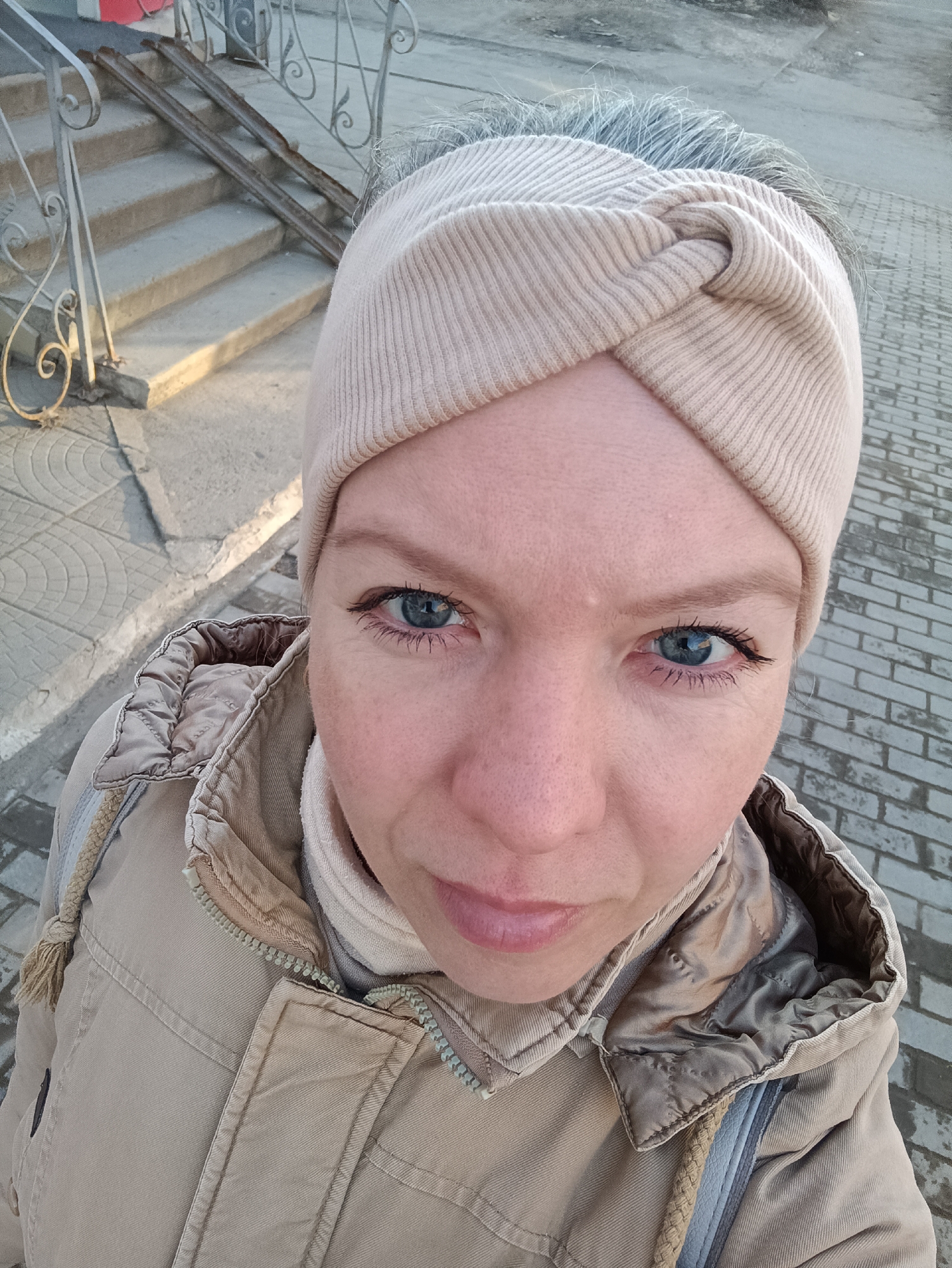 A close-up, eye-level outdoor portrait captures a woman looking directly at the camera. She has blue eyes, light skin with a few freckles, and light gray hair visible around her forehead, partially covered by a beige, ribbed knit headband with a knotted detail at the top. She is wearing a warm, beige winter jacket with a quilted lining visible around the collar and a prominent zipper. The jacket also features a drawstring on the hood.
The background shows an urban or suburban setting, possibly in Chaikovsky, Russia, as per the location hint. To the left, a set of concrete stairs with an ornate, light-colored metal railing ascends upwards. Parallel to the stairs and railing, two rusty metal rails, possibly for a ramp, are also visible. The ground is paved with a mix of light-colored patterned concrete slabs and grey brick-like pavers. The scene is illuminated by warm, directional sunlight, suggesting either early morning or late afternoon, creating soft shadows and highlighting textures. No visible text is present in the image.