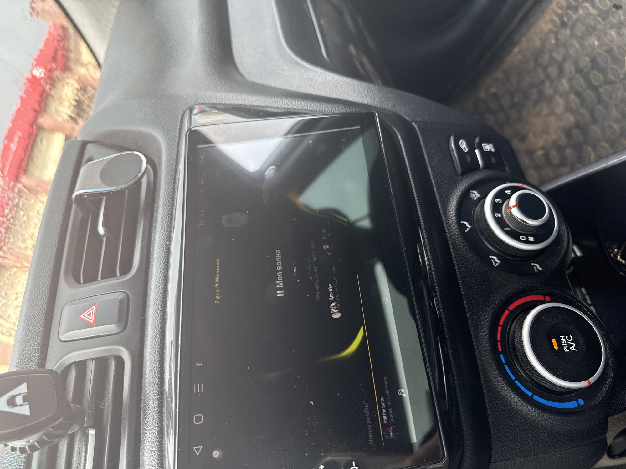 The image shows the interior of a car. The focus is on the center console, which includes a touchscreen display showing a music application, likely "Yandex Music," as indicated by the text "Яндекс Музыка" (Yandex Music). The music app displays song titles like "Моя волна" (My Wave) and "Still the Same," along with artist names and categories such as "Новые релизы" (New Releases) and "Для вас" (For You).
To the right of the screen are car climate control dials, one of which has illuminated segments indicating temperature settings, with "PUSH A/C" visible. Above the climate controls are buttons, though their function is not clear from the image. The car's dashboard appears to be made of black plastic with a textured finish.
To the left of the screen, a red hazard light button is visible within a ventilation grille. A car phone mount is attached to the left side of the dashboard. The overall setting is inside a vehicle, suggesting the activity is likely driving or a pause during a journey. The time displayed on the music app is "15:22," indicating it is mid-afternoon. The lighting suggests it is daytime. The car appears to be parked in a location with some outdoor elements visible through the windshield, which is partially obscured by what looks like water droplets or condensation, hinting at potential rain or recent washing. The overall mood is mundane, related to everyday use of a vehicle and its entertainment system.