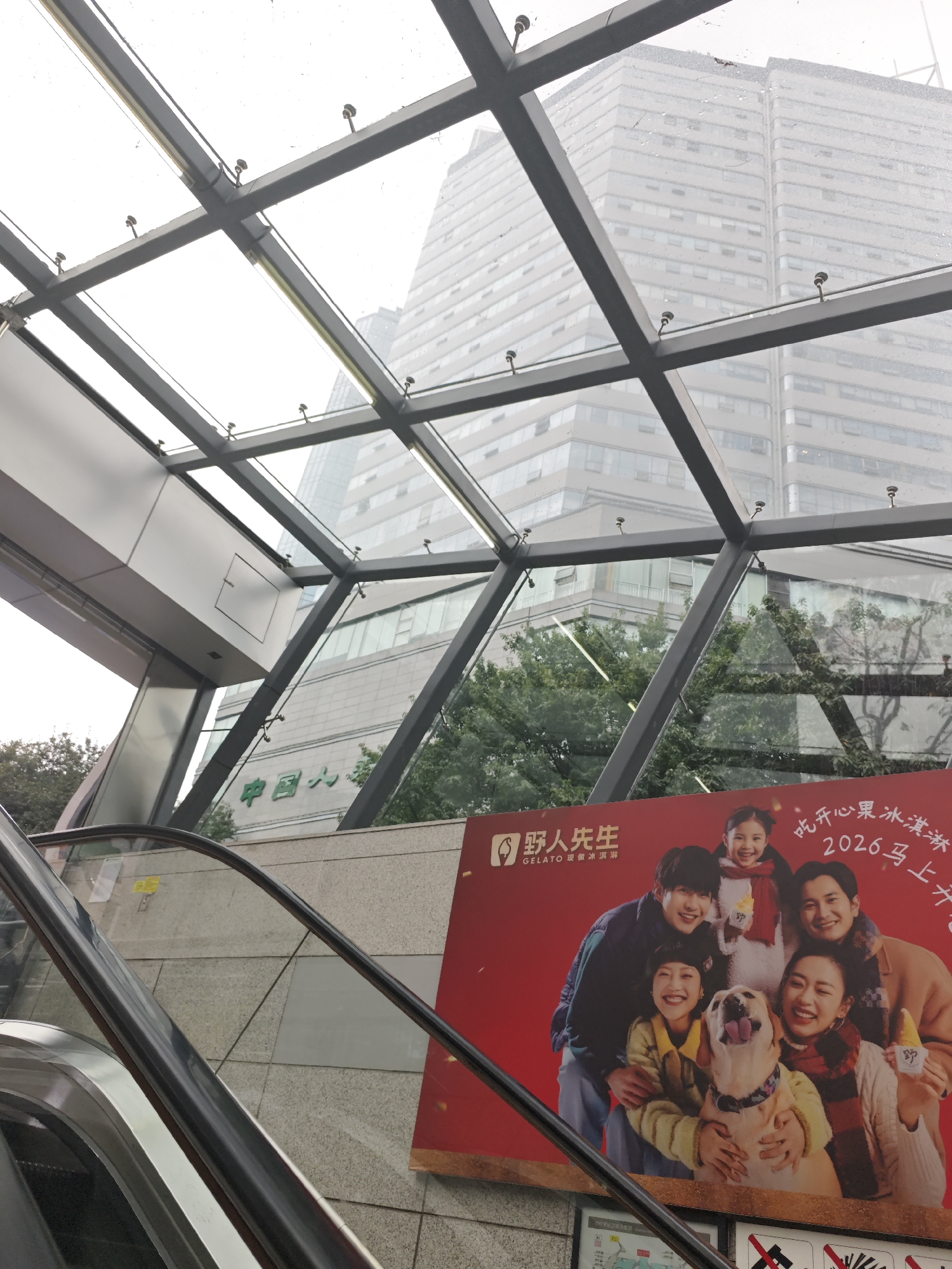 The image depicts an escalator leading up to a covered area with a steel grid and glass roof. Outside the glass roof, two tall buildings are visible in the background, appearing hazy or covered in mist. Below the glass roof and in front of the escalator, there is a large red advertisement featuring a group of people and a dog, with Chinese text and branding. The setting appears to be an urban environment, possibly at an entrance to a building or transport hub.