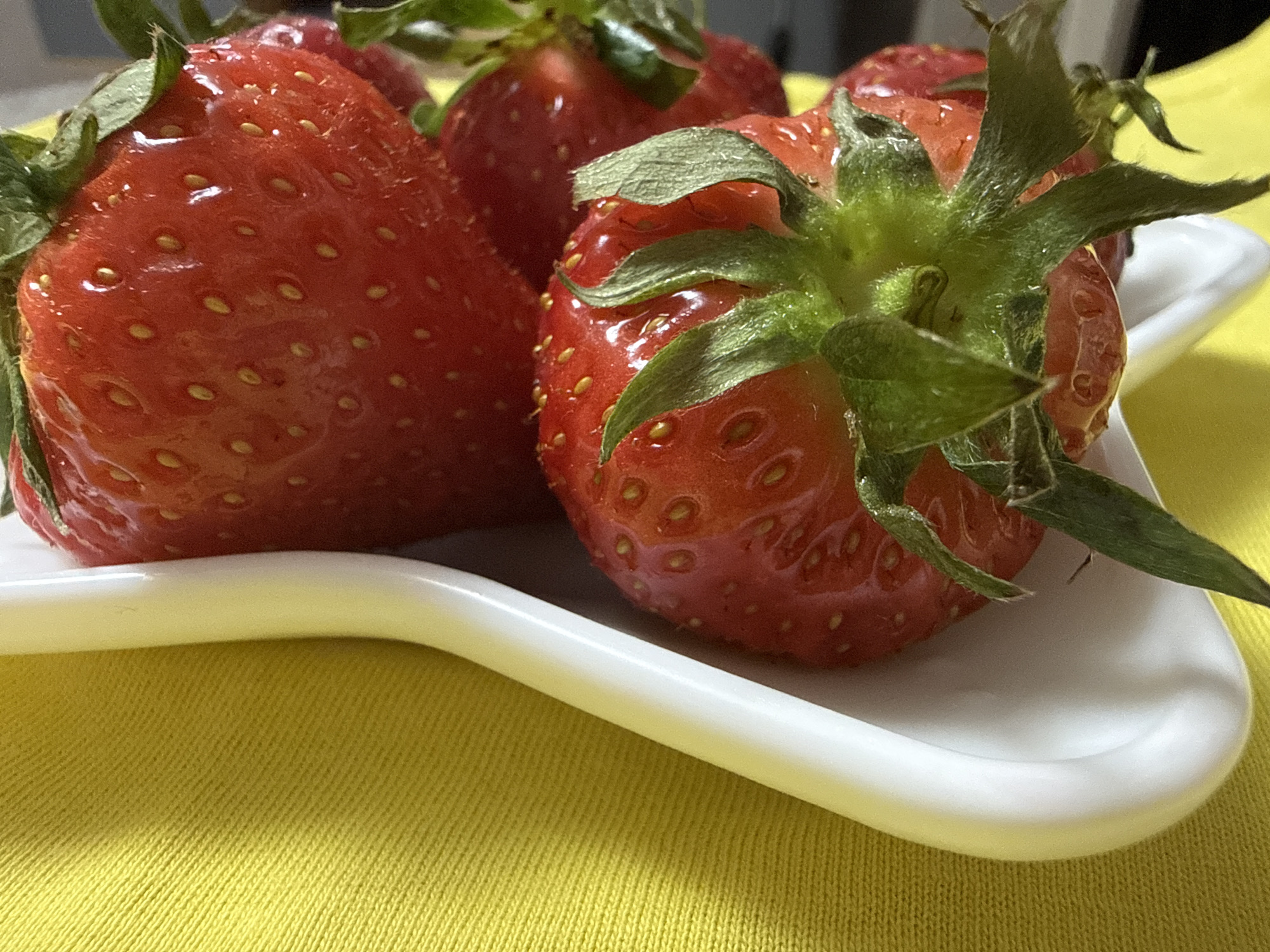 A close-up shot features a small white dish holding several fresh strawberries with green stems. The dish is placed on a vibrant yellow fabric surface. The focus is sharp on the strawberries, highlighting their red color, texture, and the small seeds on their surface. There is no indication of the location being Chaikovsky, Russia, in the image. The scene depicts a still-life arrangement of strawberries on a dish.