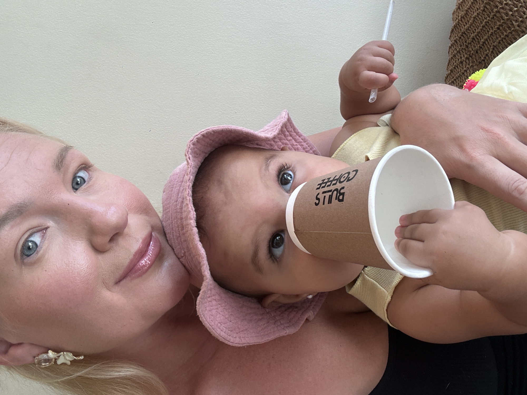 A Caucasian woman and a baby are the primary subjects of the image. The woman is fair-skinned with blonde hair and is wearing earrings. The baby, also fair-skinned, is wearing a pink bonnet and a yellow onesie. The baby is holding a paper cup with the words "Bulls Coffee" printed on it. The baby is also holding a straw in their other hand. The background appears to be a plain wall and a wicker object. There is also a yellow object with pink and green colors in the background. Given the "Bulls Coffee" reference, and the instruction to include the location of North Kuta, Indonesia, it is likely the image was taken in or near a Bulls Coffee location in that area.