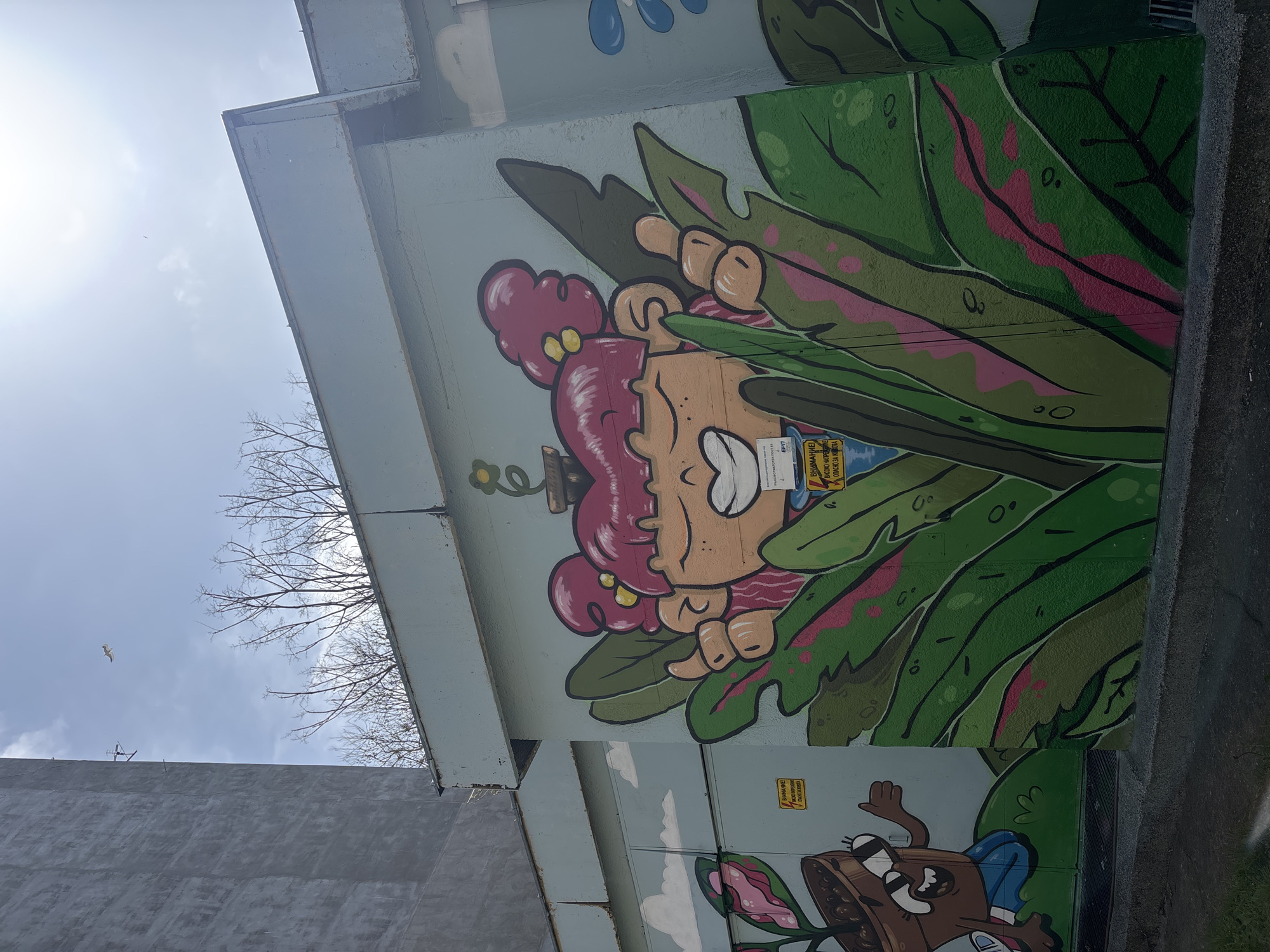 The image captures a section of a concrete structure, possibly a bridge or overpass, adorned with vibrant graffiti art in Varna, Bulgaria. The dominant mural features a cartoonish girl with pink hair peeking out from behind large green leaves, resembling a whimsical jungle scene. The artwork is detailed with bold outlines and shading. Below this, another character in a brown suit, also in a cartoon style, is partially visible. Warning signs in Cyrillic are placed near both characters. The architectural setting is a gray, industrial-looking concrete, contrasting with the colorful art. The sky is visible with a bright sun, bare tree branches, and a single bird flying. The photo appears to be taken from a low angle, emphasizing the height and scale of the structure.