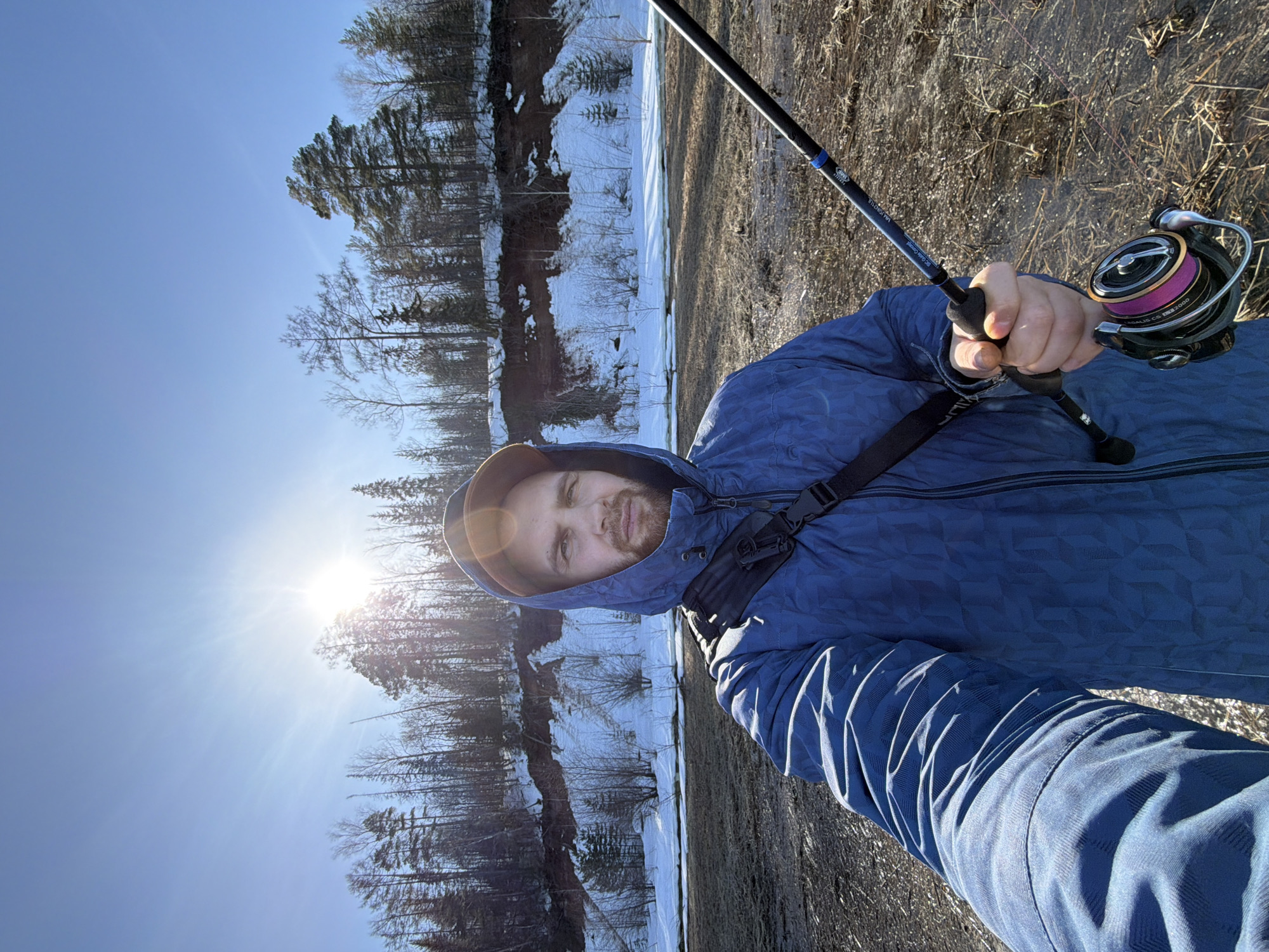 A male individual, appearing to be in his 20s or 30s with a beard, is depicted in a selfie taken outdoors in an Unknown, Unknown location. He wears a blue hooded jacket with a geometric pattern, the hood pulled up over a brown cap, and a black strap across his right shoulder. His right hand grasps a fishing rod, specifically a black model marked "ML-K102UL SG-Guide Concept", fitted with a silver and black spinning reel spooled with pink line, marked "LT2000". The foreground consists of uneven, muddy ground interspersed with dry grass and patches of standing water or ice. A body of water or partially melted ice separates the foreground from a snow-covered earthen bank. This bank is topped by a dense forest composed of bare deciduous trees and evergreen conifers. The sky is clear blue with a bright sun positioned high, causing a prominent lens flare and illuminating the scene, indicating a sunny day during a transitional season, likely late winter or early spring.