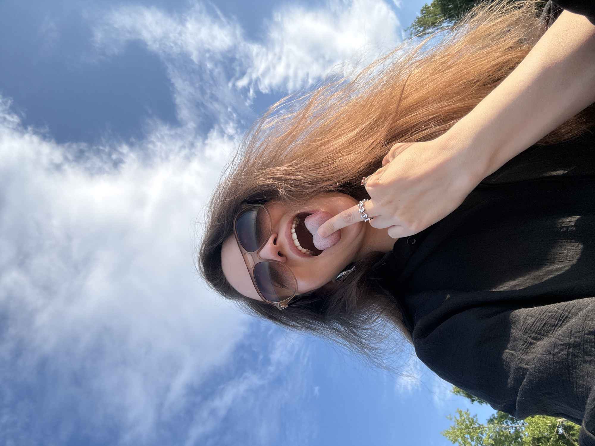 A woman with long, brown hair and sunglasses sticks her tongue out while holding her finger to her mouth and wearing a black shirt and rings. The background shows a blue sky with clouds and some trees. The location is the city of Chaikovsky, Russia.