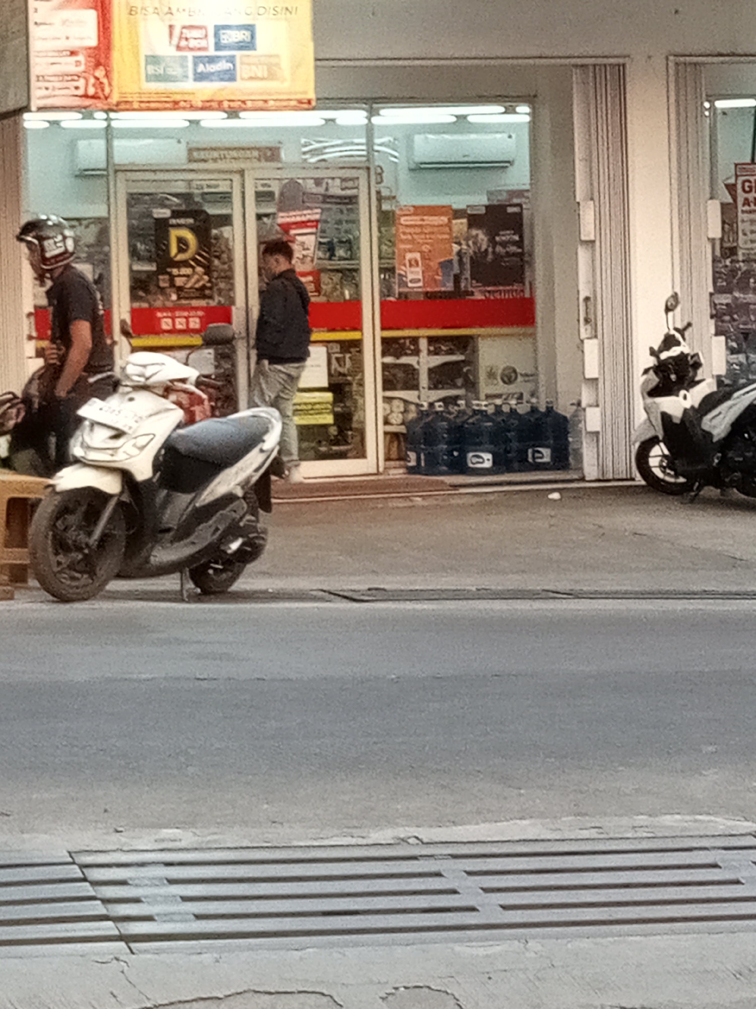 The image depicts a street scene in front of a convenience store, possibly located in Sariwangi, Indonesia. The store's facade features glass doors and shutters, and a prominent banner displaying various bank logos is visible above the entrance. Inside, merchandise and an air conditioning unit can be seen. Outside the store, two men are present. One man, wearing a black t-shirt and a helmet, stands near a parked white scooter. The other man, in a dark jacket and light-colored pants, stands further inside the store's entrance. A row of large water jugs is stacked on the pavement near the store entrance. Another white scooter is parked to the right of the store. The foreground shows a gray asphalt road with a metal drainage grate. The lighting suggests it is daytime.