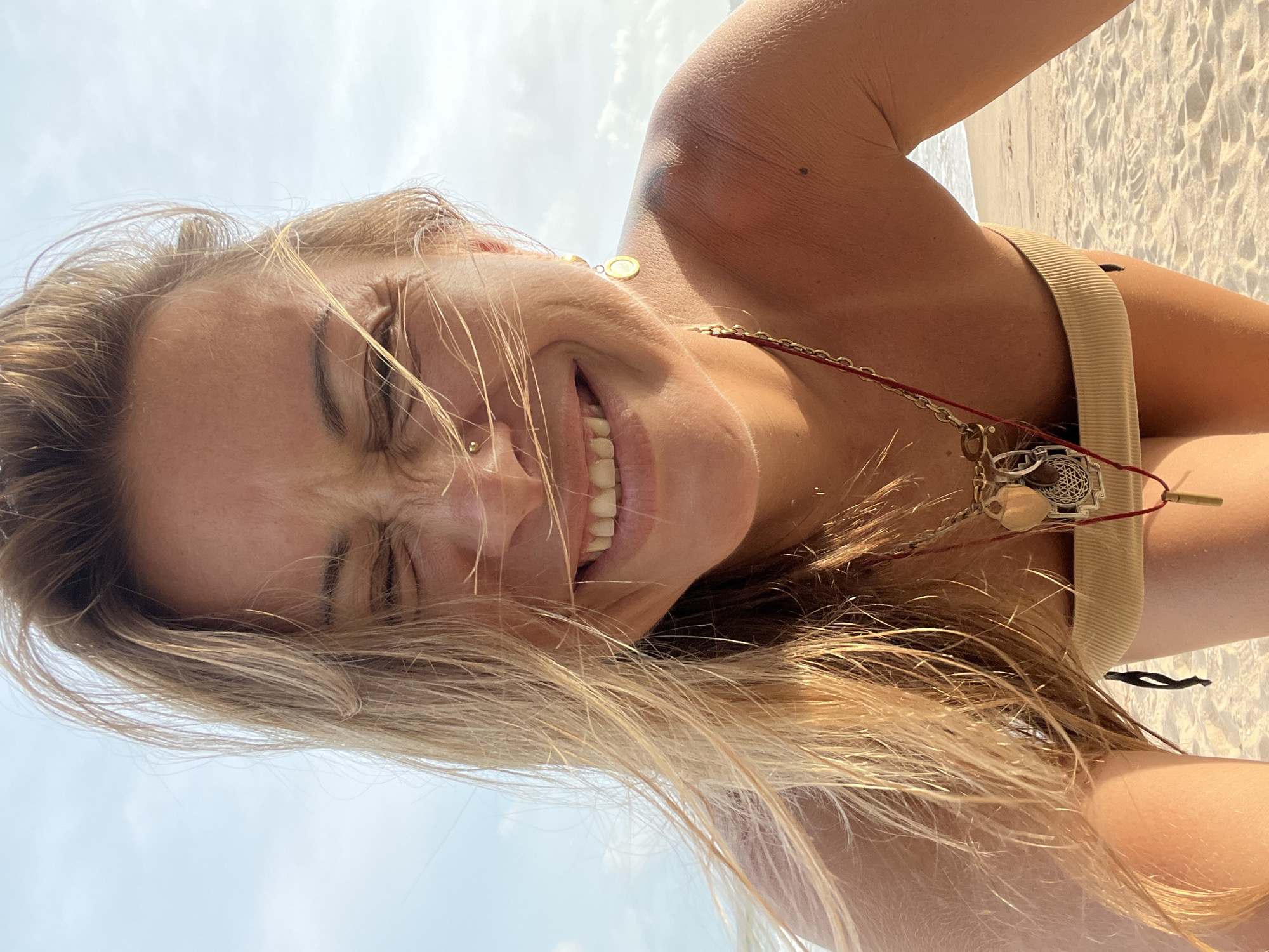A close-up, vertically oriented selfie shows a young woman with blonde hair against a bright sky and beach background. Her eyes are squinted, and she is smiling widely, with her teeth visible. A nose ring and an earring are visible. She is wearing a tan bikini top and a necklace with several charms. Her hair is blowing in the wind. The beach sand, with footprints visible, and a hint of the ocean are in the background, suggesting a sunny day. The overall mood is joyful and relaxed, indicative of a vacation or leisure activity at the beach. The setting is identified as Peleana, Sri Lanka. There is no visible text in the image.