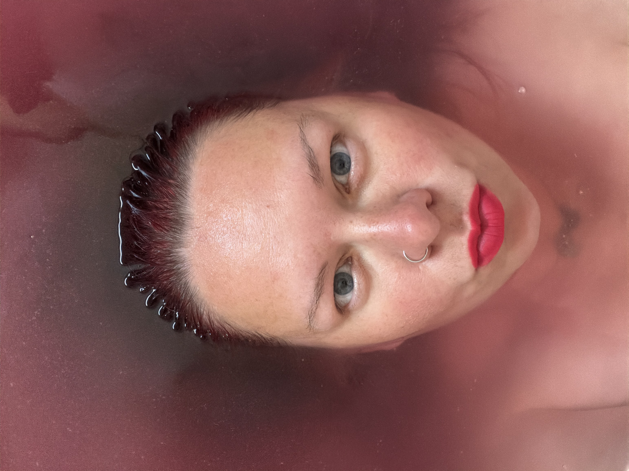 A woman with red hair and bright red lipstick is submerged in a body of red-tinted water. The woman is facing up toward the viewer, with only her face and the edge of her hairline visible above the water. The woman has fair skin, blue eyes, and a nose ring. A small water droplet clings to the side of her neck. The photo was taken at an indeterminate location in Chaikovsky, Russia.