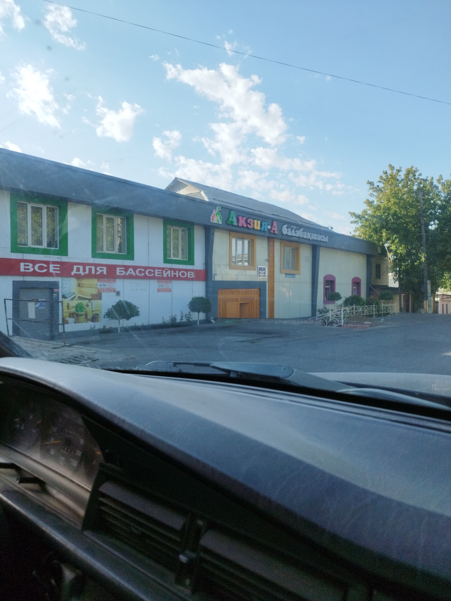 The image depicts a storefront for "Акция-А" (Aktsiya-A) which translates to "Action-A", a store specializing in "ВСЕ ДЛЯ БАССЕЙНОВ" (VSE DLYA BASSEYNOV), meaning "Everything for pools". The store is a commercial building with a green and white facade. It has multiple windows with green frames and white interiors. A prominent red banner with the text "ВСЕ ДЛЯ БАССЕЙНОВ" runs across the lower part of the building's facade. To the right, another sign reads "Акция-А Балакщасы" (Aktsiya-A Balakshasy), suggesting it's a branch of the store. The building is set in an urban or semi-urban environment with trees and a cloudy sky visible. The foreground shows the interior of a car, indicating the photo was taken from inside a vehicle. The image appears to be taken in Shymkent, Kazakhstan, based on the store name and the context provided.