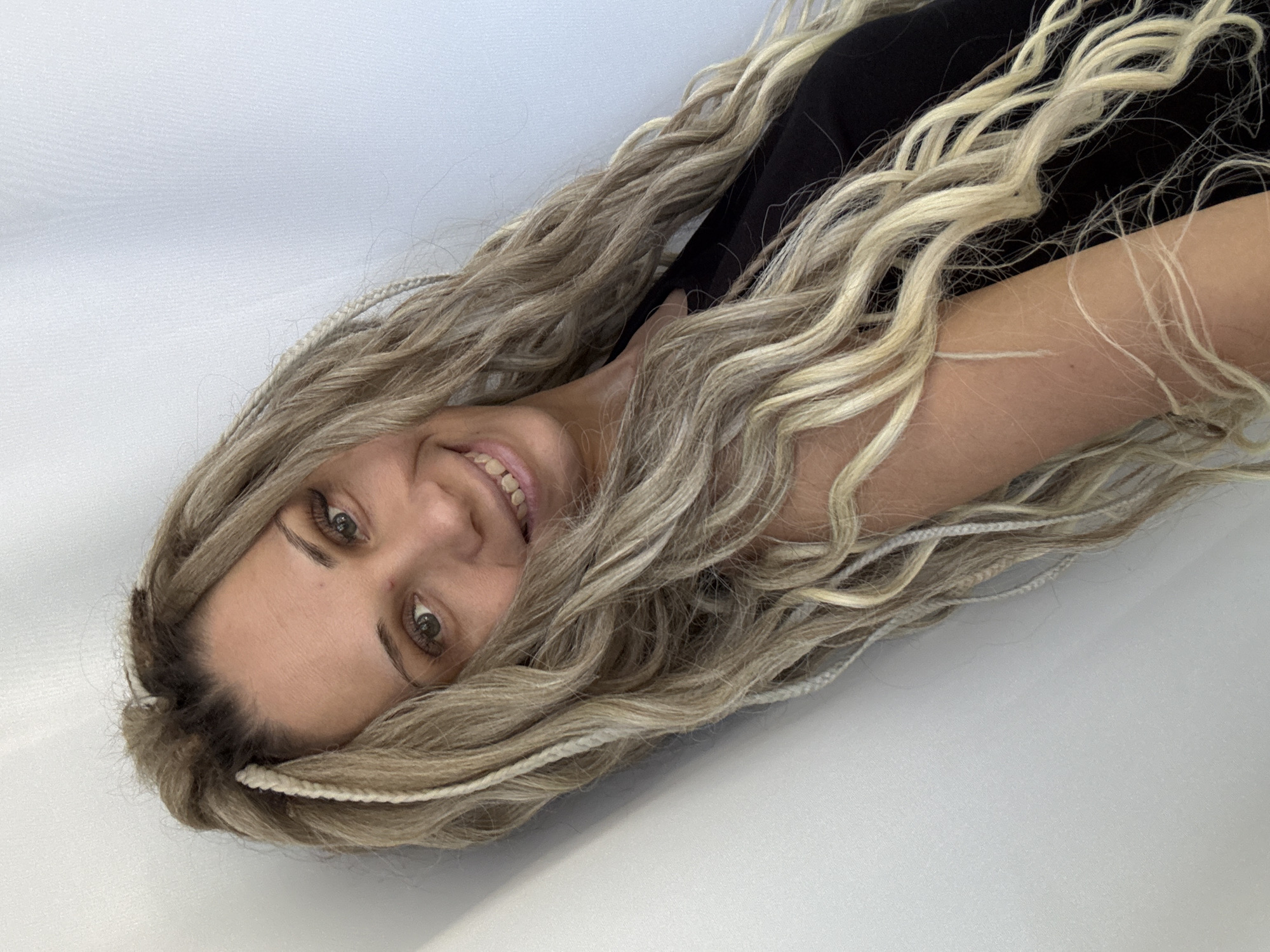 A light-skinned woman with long blonde braided hair is smiling at the viewer. She is wearing a black top. The background is a light-colored surface. The location is not discernible from the image.