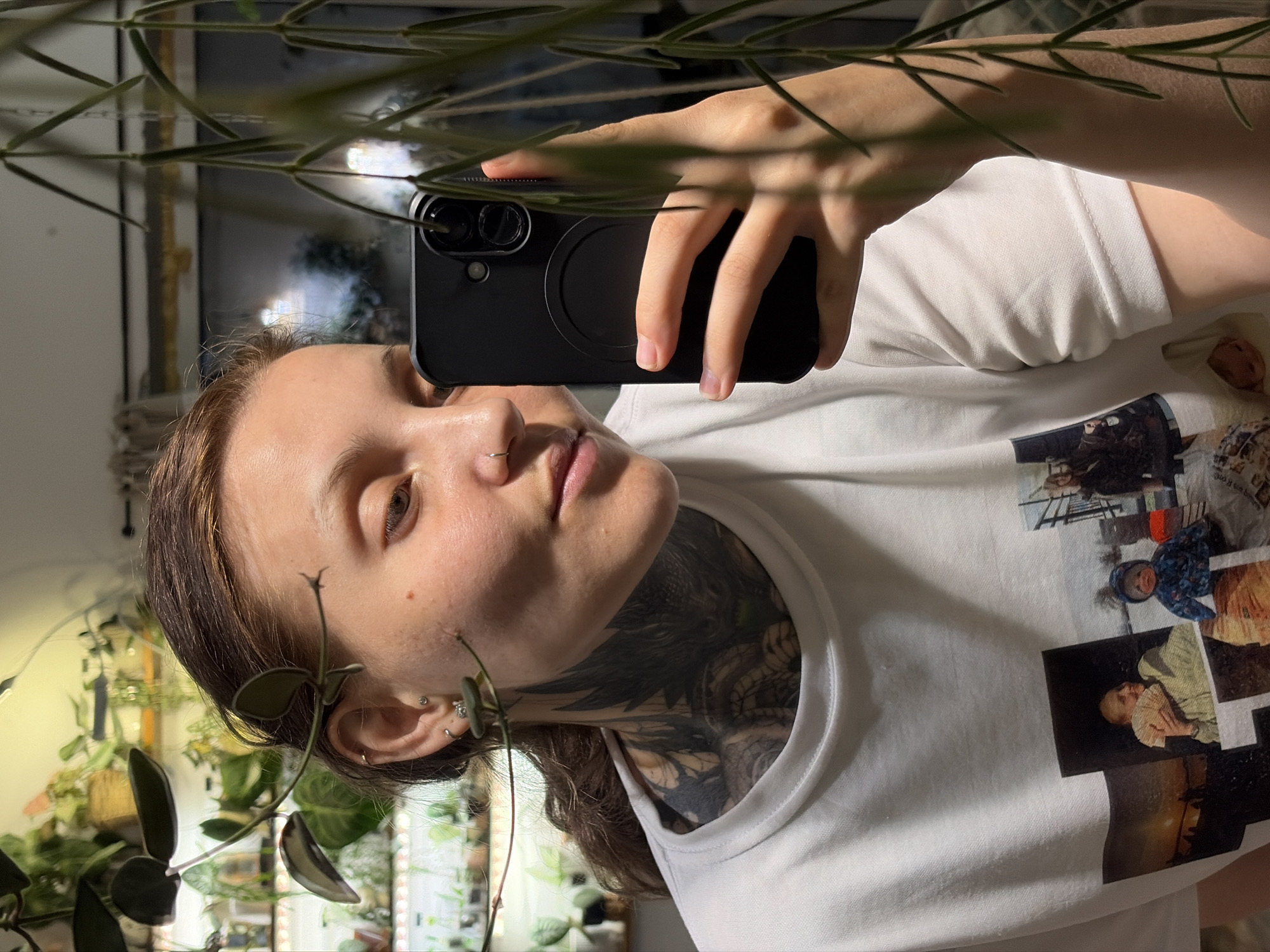 The image is a selfie taken by a young woman with light brown hair, who appears to be indoors. She is wearing a white t-shirt with several printed photographs on it, one of which depicts a baby in a blue hooded jacket. She has a nose ring and multiple ear piercings, and a large, dark tattoo covering her neck and chest. She is holding a black smartphone in her right hand, in the process of taking the photo. In the background, numerous plants are visible, suggesting an indoor garden or a room with many houseplants. The lighting is dim, with some artificial light sources present, casting shadows. The woman's expression is neutral, and she is looking towards her phone screen. No specific city or geographical location is identifiable in the image, and therefore no information can be provided about the city of Malyi Bukor, Russia.