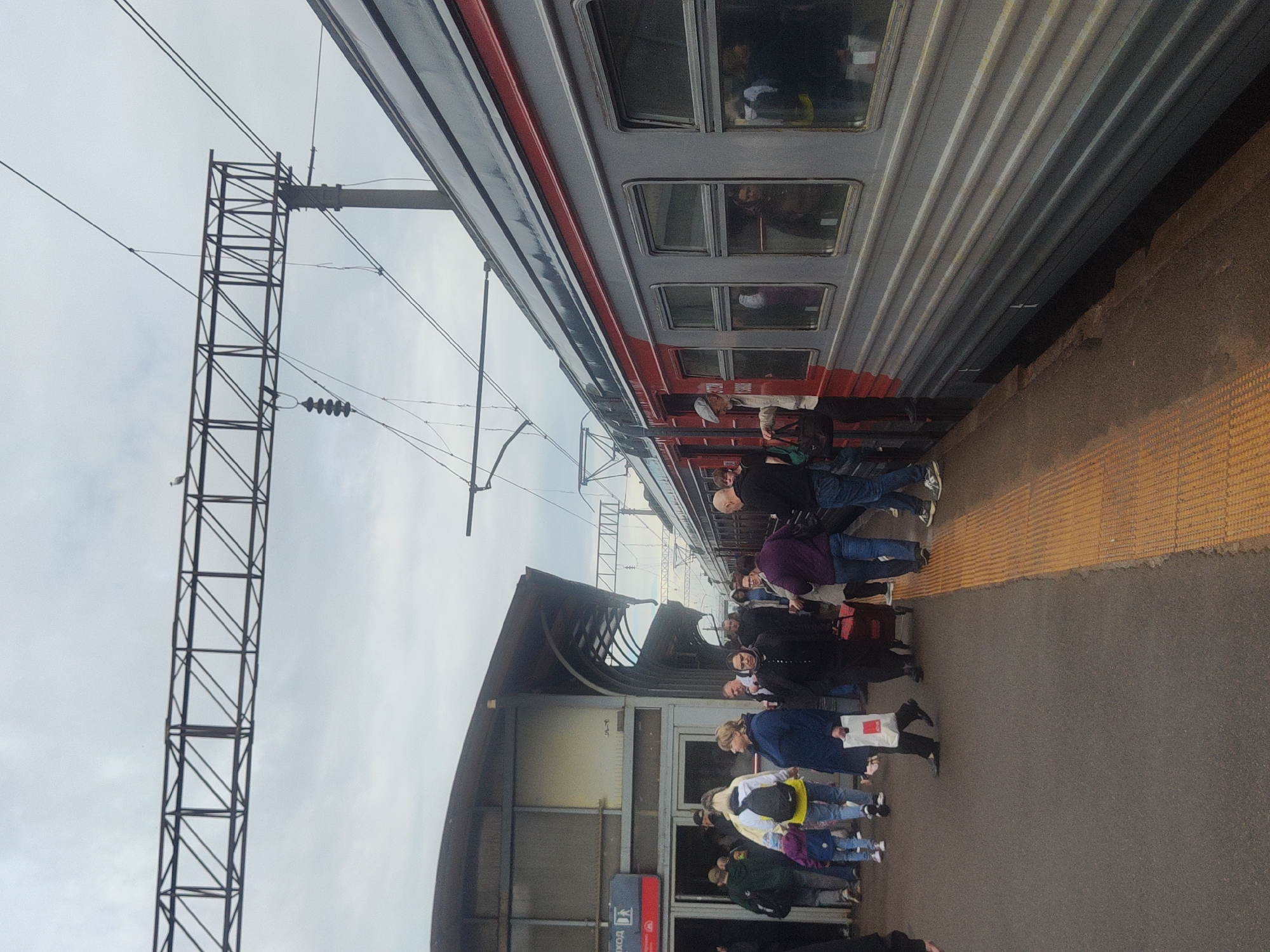 The image depicts a train platform scene. People are standing alongside a parked train, likely boarding or disembarking. A metal structure with support wires is visible on the left. The platform has tactile paving. A building with windows and a covered walkway are visible in the background. The sign on the building is written in Russian, indicating the location is likely Saint Petersburg, Russia.