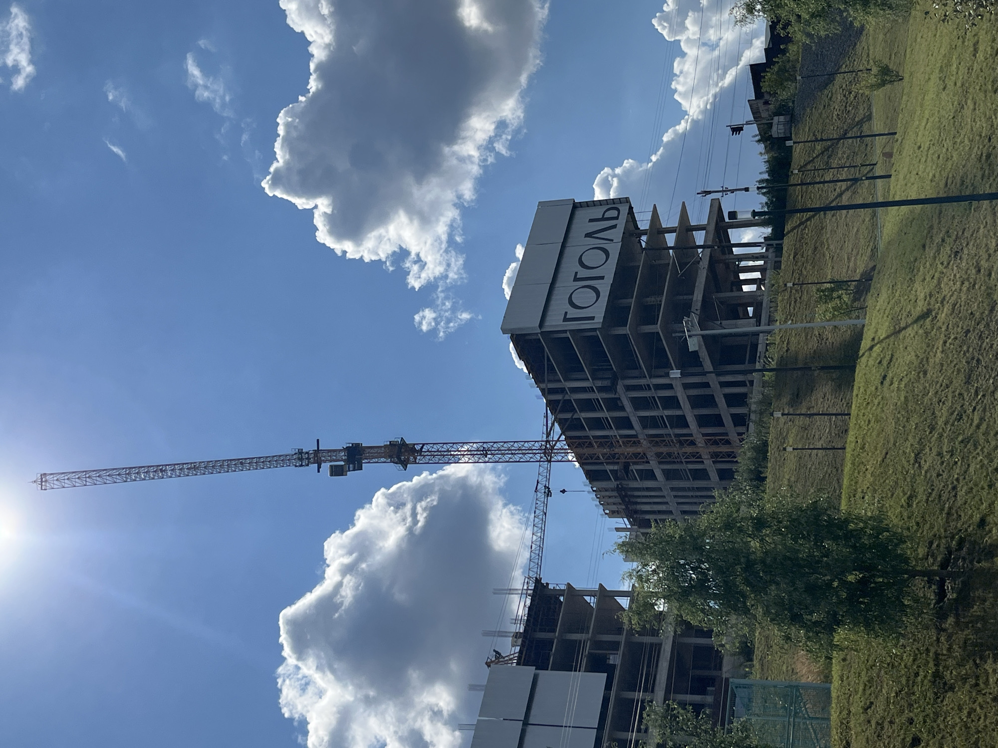 The image shows a construction site for a building with the word "ГОГОЛЬ" (GOGOL) in large black letters on its facade. Two construction cranes are visible, one tall and one horizontal, suggesting ongoing construction. The building appears to be made of concrete and is in an unfinished state, with multiple floors and exposed rebar.
The location is the GogoL residential complex in Perm, Russia. The scene is set against a bright blue sky with fluffy white clouds. In the foreground, there is a grassy slope with some trees and lampposts. The overall impression is of a modern urban development project in progress under a sunny sky.