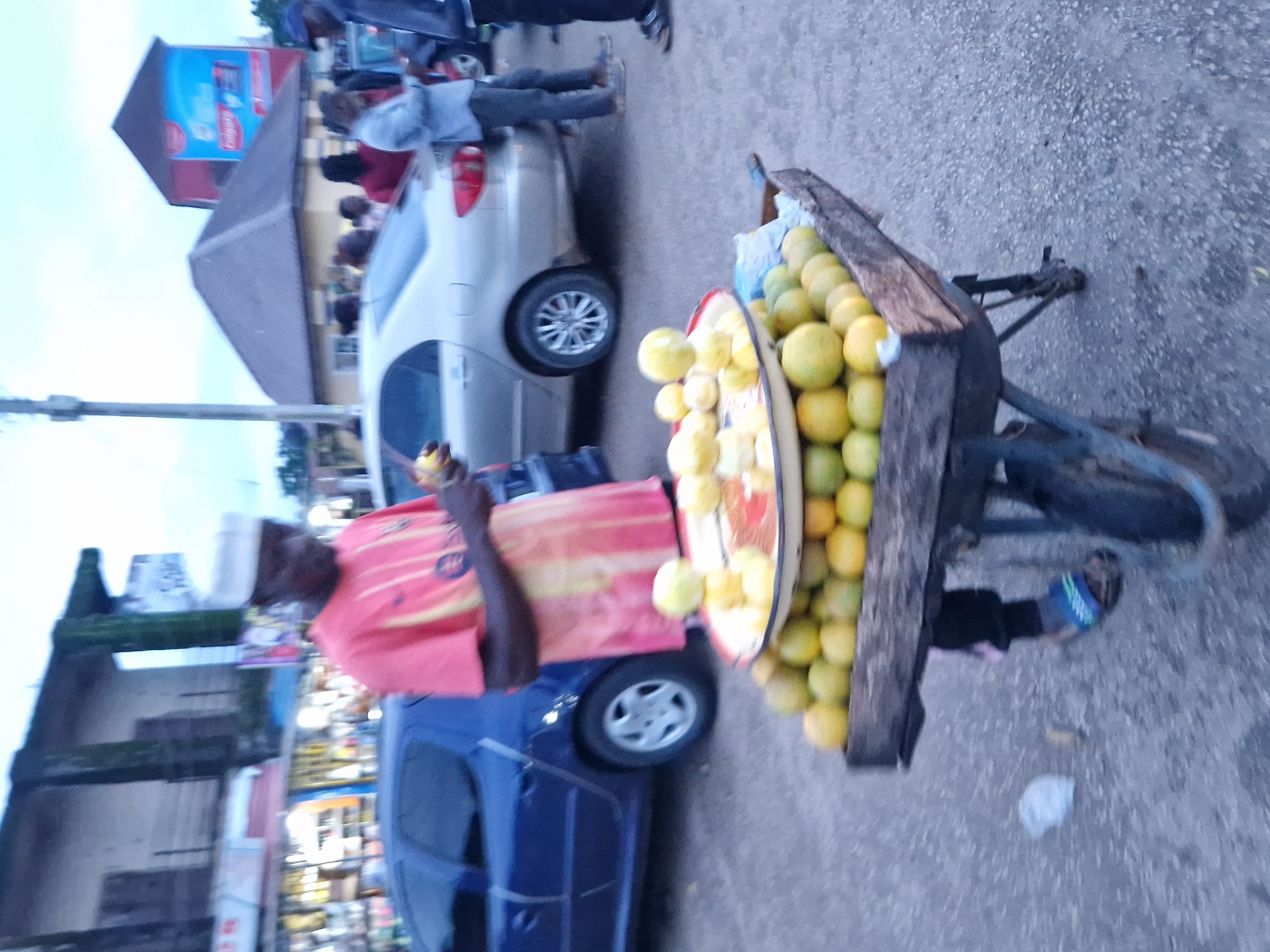 The image shows a street scene in Kabong, Nigeria. A man wearing a red and yellow striped shirt and a white cap is standing behind a fruit cart laden with oranges and lemons. He is holding a piece of fruit in his hand, possibly in the process of selling it. The cart is a wheelbarrow with a wooden frame. In the background, there are cars, buildings, and other people. One building in the background has a large blue and red sign for Colgate toothpaste. The sky is overcast.