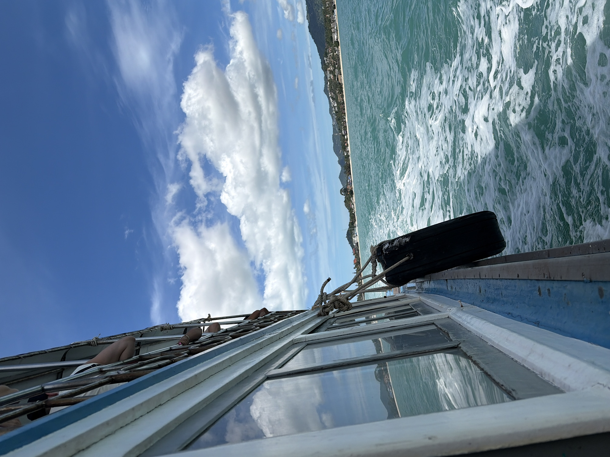 This media file depicts a view from a boat at sea, looking towards a coastline. The scene is oriented vertically, suggesting the photo was taken while the boat was in motion or tilted.
The subjects include a section of the boat's deck, with visible windows reflecting the sky and water. On the deck, there are glimpses of people, their legs and feet casually resting, suggesting they are passengers enjoying the journey. A black fender, likely for cushioning the boat against a dock or other vessels, is secured to the side of the boat with thick ropes. The water is a turquoise hue, with white foam and ripples indicating movement, possibly a wake from the boat or waves.
In the distance, a long stretch of coastline is visible, lined with buildings that suggest a populated area, possibly a town or resort. Mountains rise behind the buildings, adding to the scenic backdrop. The sky is a vibrant blue, dotted with fluffy white cumulus clouds, indicative of a bright, sunny day.
The overall impression is one of a relaxed boat trip, likely during daylight hours given the clear sky and strong sunlight casting reflections. The combination of the sea, coastline, and clear sky points to a tropical or coastal location. The mention of Baan Bang Rak, Thailand, provides specific location context, suggesting the boat is traveling in or near this area. There is no visible text in the image.