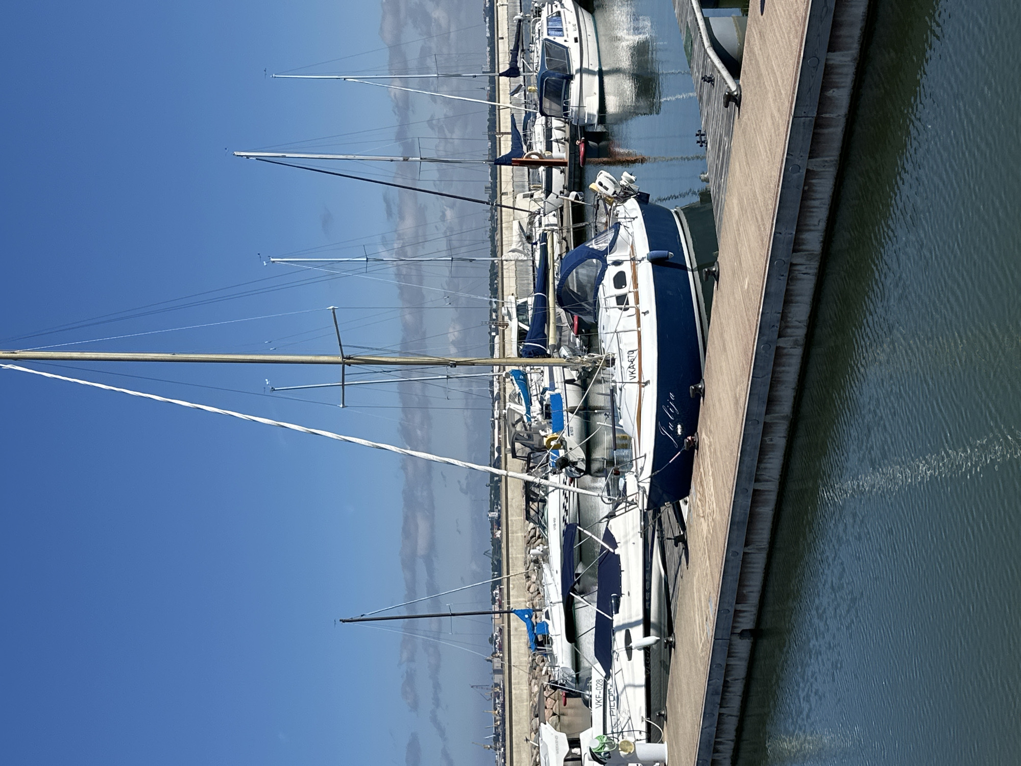 The image shows a marina in Tallinn, Estonia, on a clear day. Several sailboats and a motorboat are docked along a wooden pier. The main subject is a blue and white sailboat in the foreground with the name "Julija" visible on its hull. In the background, more boats are moored, and a concrete barrier separates the marina from the city, with industrial structures and cranes visible in the distance. The water is calm with reflections of the sky and boats. No people are visible in the image.