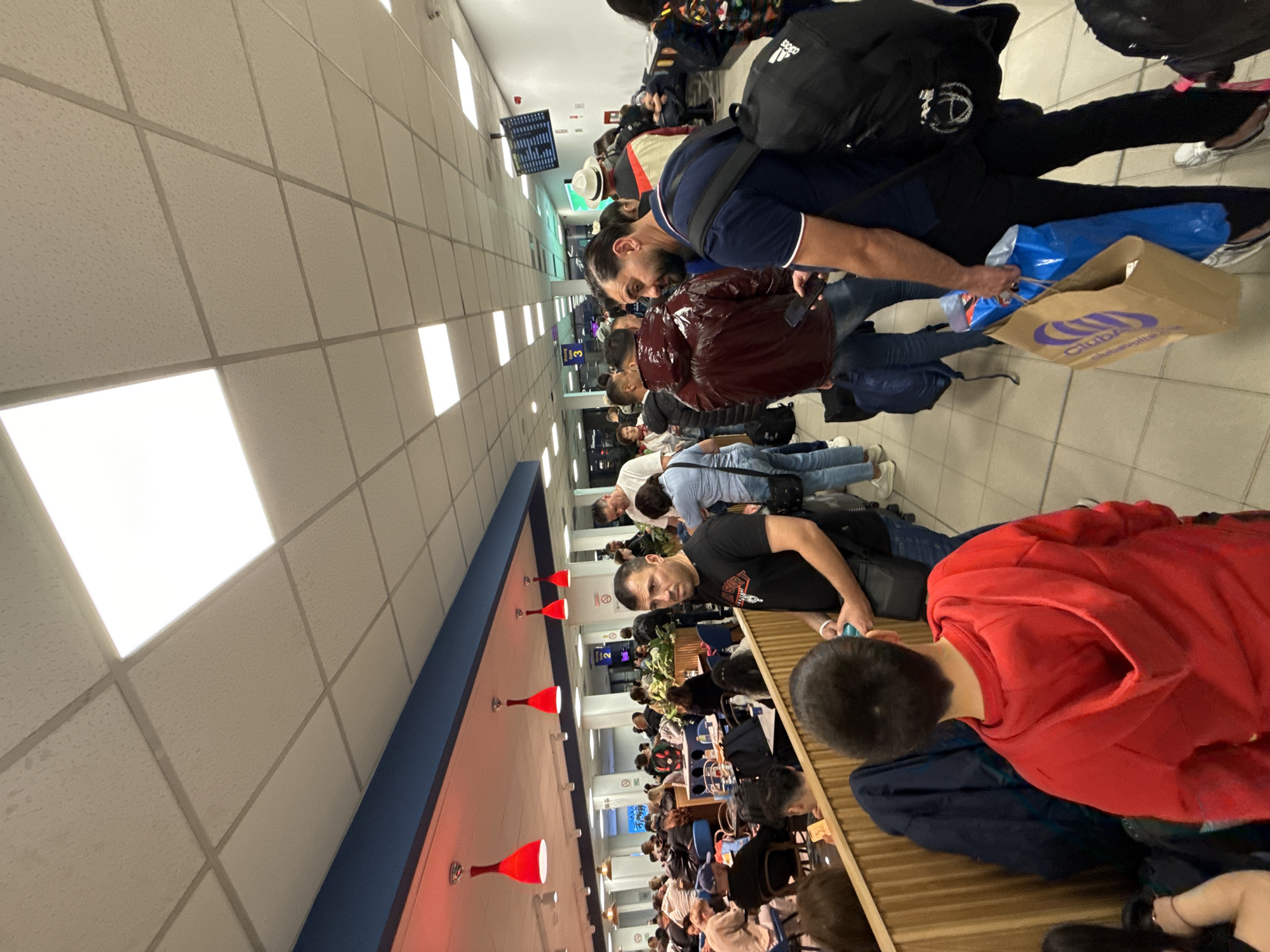 The vertically oriented image depicts a crowded indoor public space, identified as likely an airport terminal in Sofia, Bulgaria. Numerous individuals, mostly adults, are either standing in what appears to be a queue or waiting area, or seated at tables and benches further into the background. Many people are equipped with various pieces of luggage, including backpacks and carry-on bags, indicative of travel.
In the mid-ground, a man in a dark polo shirt is seen holding a blue plastic bag and a brown paper bag prominently displaying the "Club Airways" logo. Another person nearby carries a black backpack with the "adidas" logo. Further into the background, a person wears a distinctive red hooded top while seated. Many individuals are engaged with their mobile phones.
The ceiling features a grid of light-colored acoustic tiles with integrated rectangular fluorescent light panels, providing bright illumination. On the right side of the frame, a section with red, bell-shaped pendant lights hangs over an area furnished with tables and chairs, possibly a cafe or food court. In the distance, a digital display resembling a flight information board is visible, along with a partial sign that might read "Gate 3". The scene conveys an atmosphere of transit and waiting, typical of a busy travel hub. The time of day appears to be evening or night, given the reliance on artificial lighting.