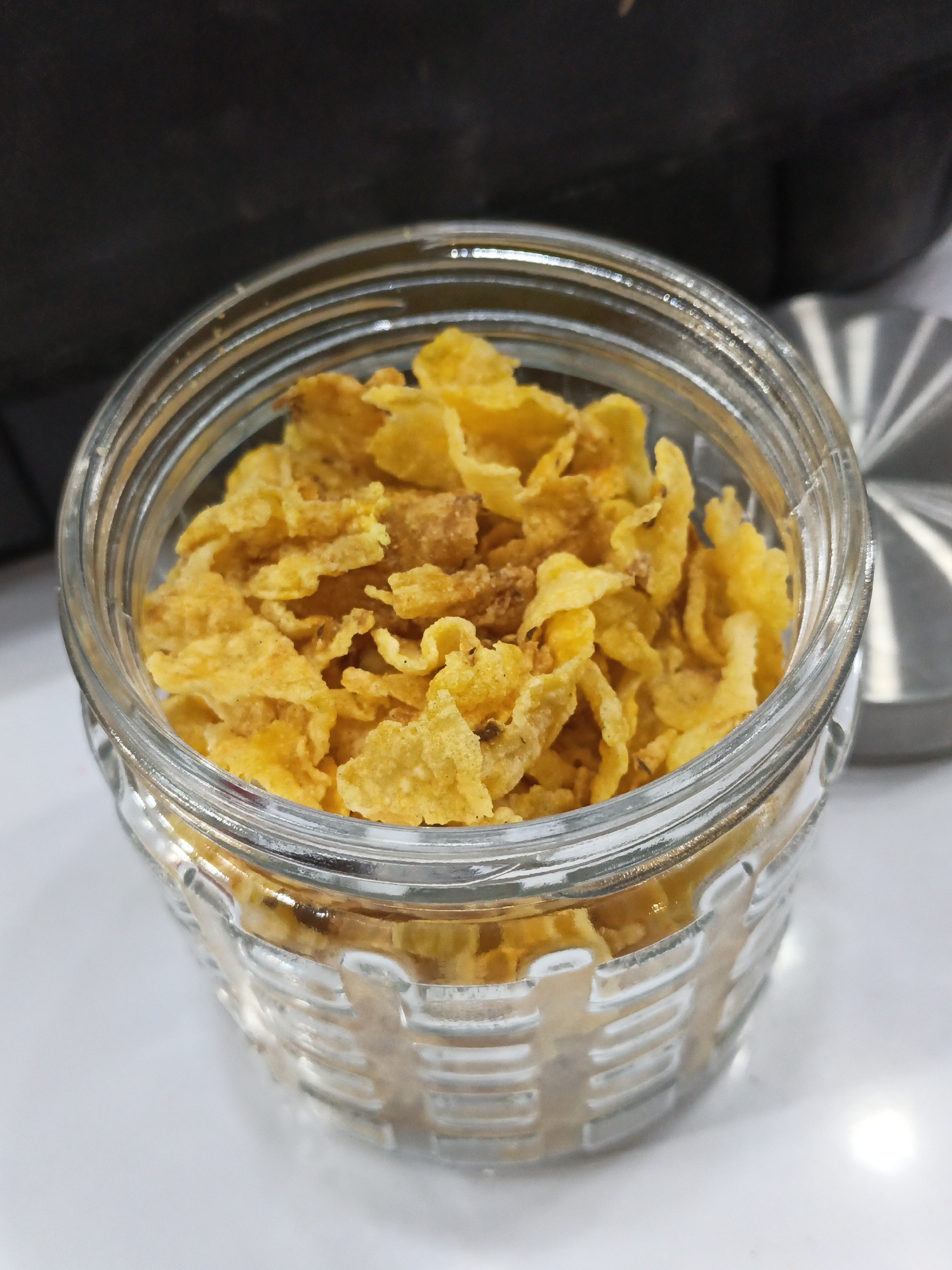 A glass jar filled with cornflakes sits on a white surface. The jar is transparent and ribbed, allowing the yellow cornflakes to be seen inside. In the background, a dark, textured object is visible, possibly a kitchen counter or appliance. To the right of the jar, a metal lid with a ridged edge is partially visible. The lighting suggests an indoor setting. This image does not contain any elements that would allow for the determination of its location in Karangpakel, Indonesia.
