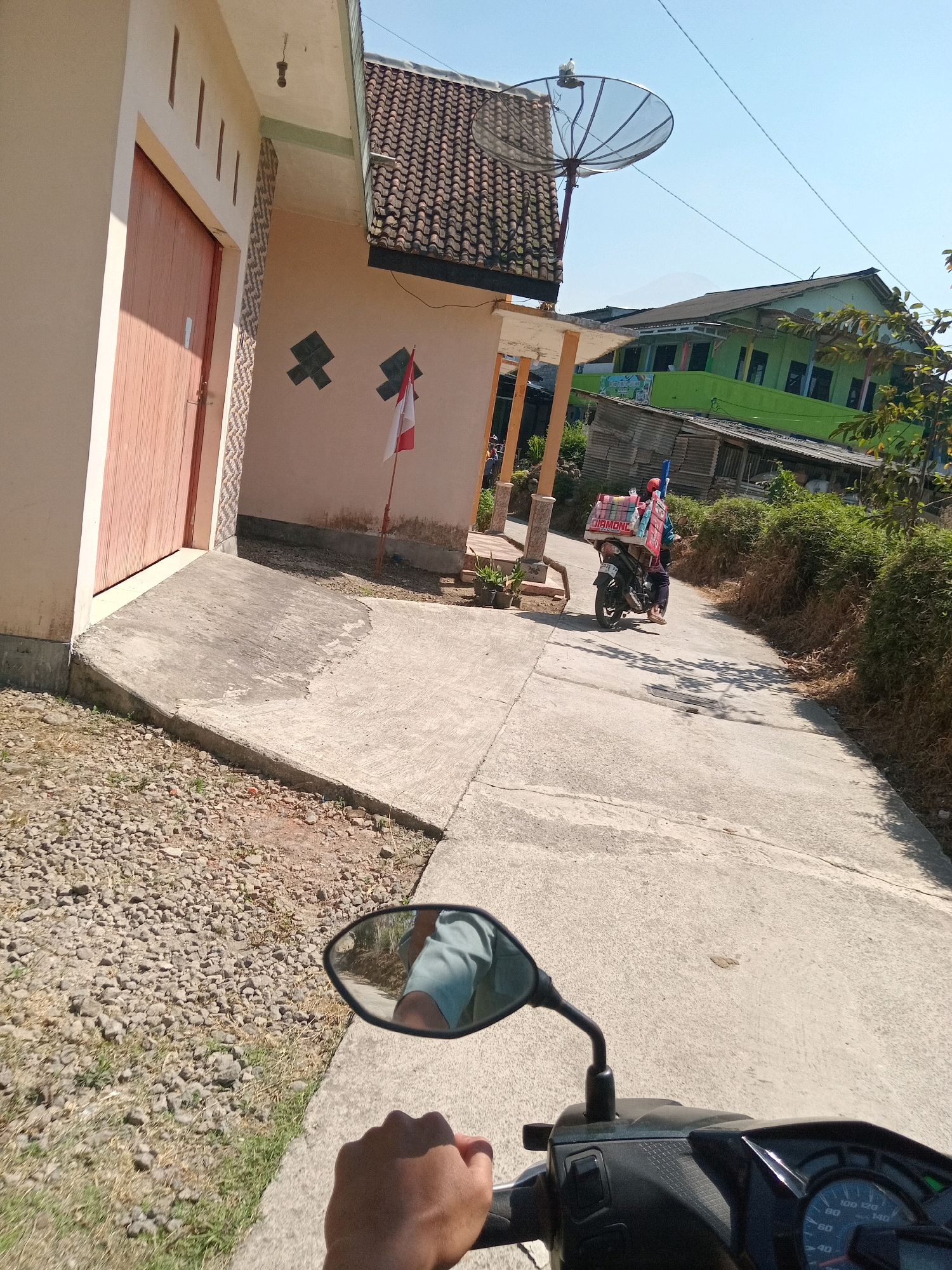 The image depicts a sunny outdoor scene in Maduretno, Indonesia, with a paved road leading uphill. In the foreground, a person's hand is visible on the handlebar of a motorcycle, with a side mirror reflecting the rider's arm and a portion of a light blue shirt. The motorcycle is moving away from the viewer.
To the left of the road, there is a light-colored building with a reddish-brown wooden door and a concrete ramp leading to it. The building has a tiled roof and decorative square patterns on its wall, along with a small Indonesian flag. Adjacent to the building, a patch of gravel and dry grass is visible.
Further up the road, a person is riding a motorcycle loaded with a large white box containing red items, possibly goods for sale, and several blue poles. The rider is wearing a blue shirt and dark pants. Behind the rider, a green building with a tiled roof and a satellite dish mounted on an adjacent structure can be seen. The background shows lush green foliage and a distant mountain under a clear blue sky. The overall environment suggests a rural or semi-rural setting within the city of Maduretno.