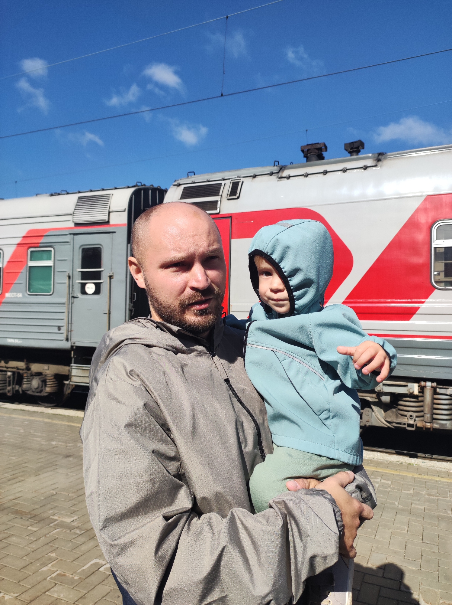 A bald, bearded man with a light brown complexion is holding a young child in his arms. The man is wearing a grey hooded jacket. The child, with blonde hair, is wearing a light blue hooded jacket and light green pants. They are standing on a paved platform in front of a train. The train is grey and red, with visible windows and doors. The sky is blue with a few white clouds. The scene appears to be a train station. The location is identified as the Kirov Railway Station in Kirov, Russia.