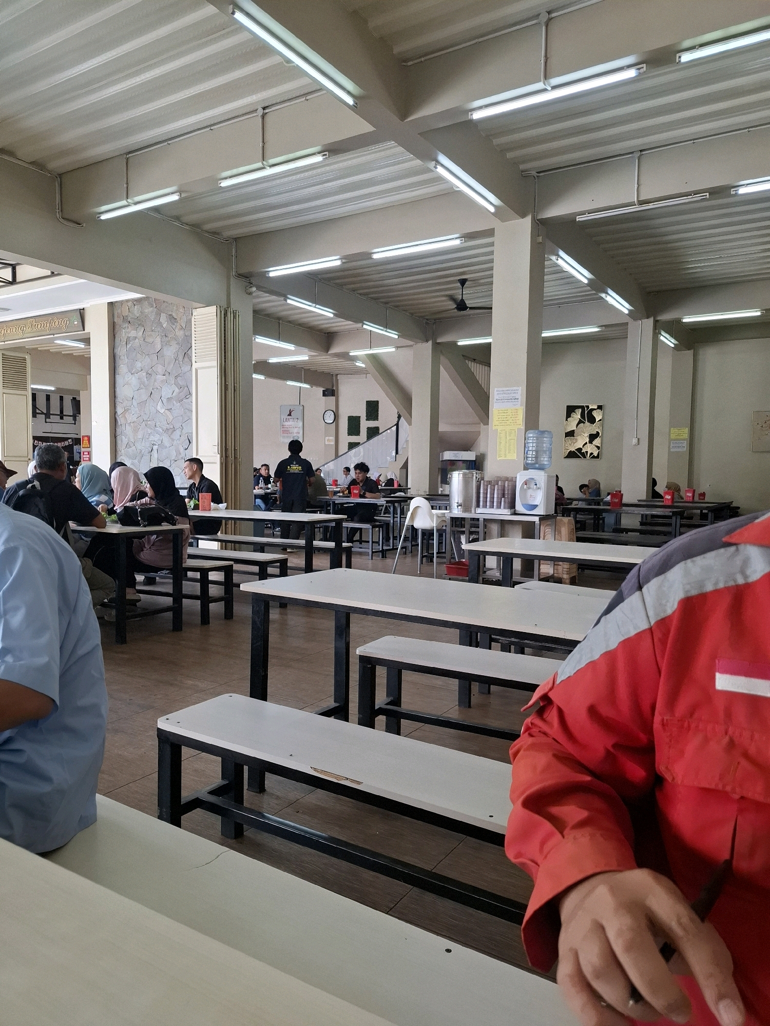 The image shows a crowded canteen with long tables and benches. Several people are seated at the tables, eating and talking. In the foreground, a person wearing a red uniform is visible, with their arm and hand resting on a table. The canteen appears to be indoors, with fluorescent lighting and a high ceiling. The overall impression is one of a busy, everyday scene in a public eating area. Based on the context provided, this scene is located in Sukabumi, Indonesia.