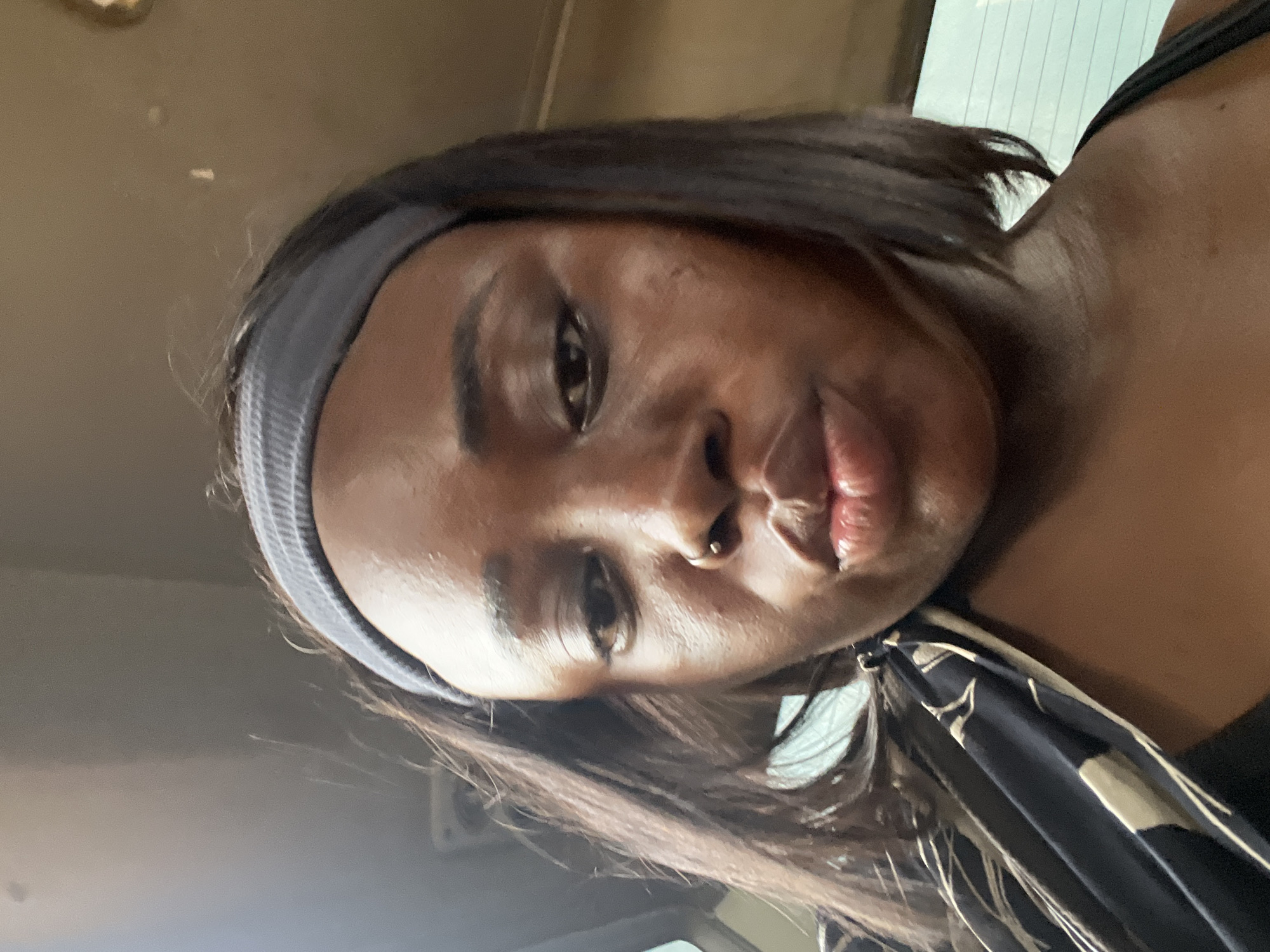 This is a close-up, overhead selfie of a young Black woman indoors, likely in a vehicle given the reflection of what appears to be a window or sunroof above and to the right of her head. She is wearing a grey headband that holds back her dark, shoulder-length hair. Her head is tilted, and she is looking directly at the camera with a neutral expression. She has a small nose ring. She is wearing a black top, and a patterned fabric, possibly a scarf or clothing, is visible draped over her shoulder. The lighting suggests it could be daytime, with natural light coming from above. The scene is otherwise nondescript, with a muted, earthy background that blends into the shadows. No text is visible in the image.