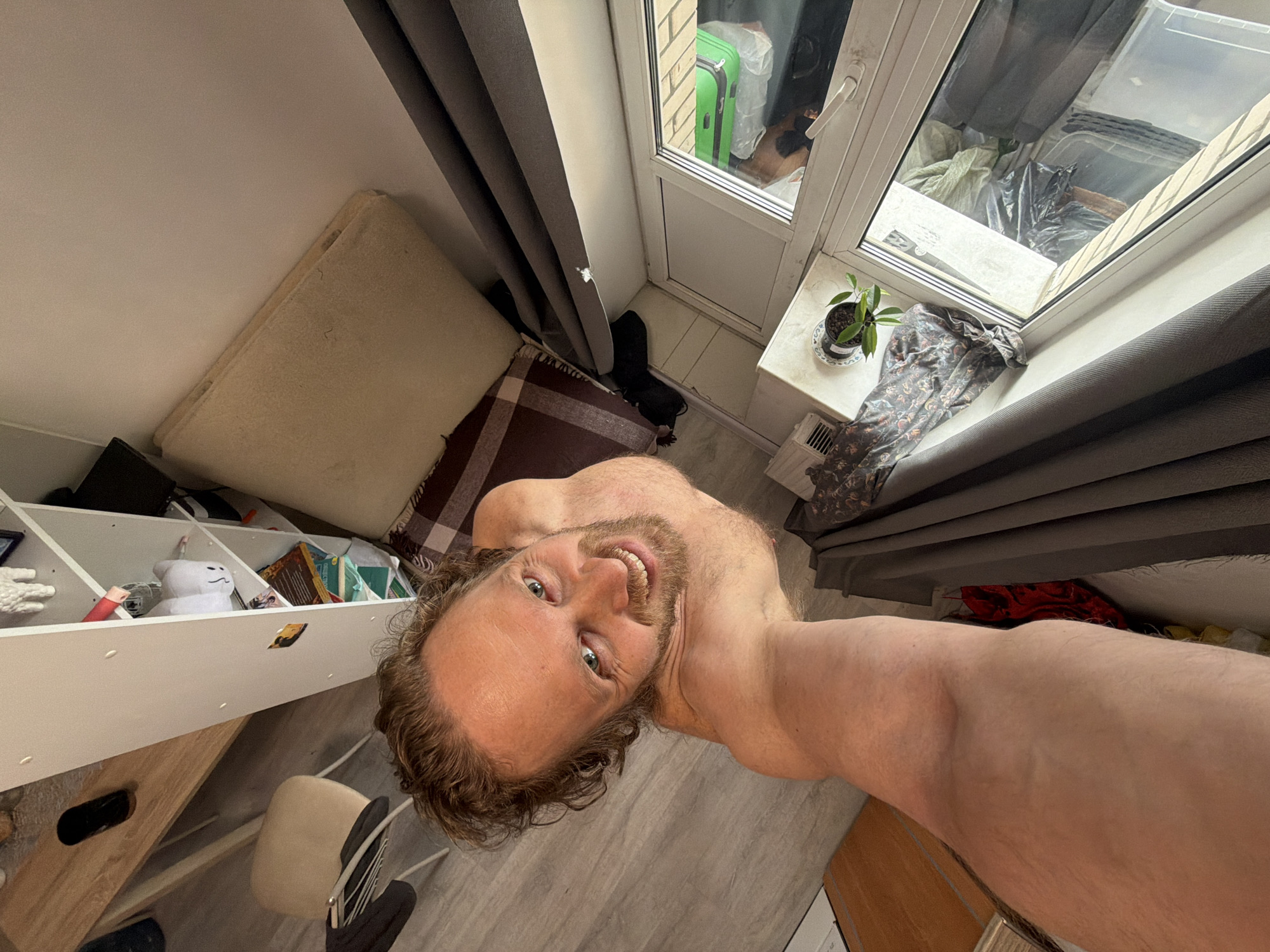 A man with light skin, curly brown hair, and a beard is taking a selfie from a low angle, facing the camera. He is shirtless. Behind him, there is a window with gray curtains. Outside the window, there is a glimpse of a green suitcase and other items, suggesting a balcony or outdoor area. A plant sits on the windowsill. There is a mattress leaning against the wall next to the window, with a brown and white checkered blanket underneath it. To the left, there is a white shelving unit filled with various objects, including a white plush toy. A grey chair is located near the bottom left. The location is Saint Petersburg, Russia, as inferred by the window frame style, typical of apartment buildings in the city.