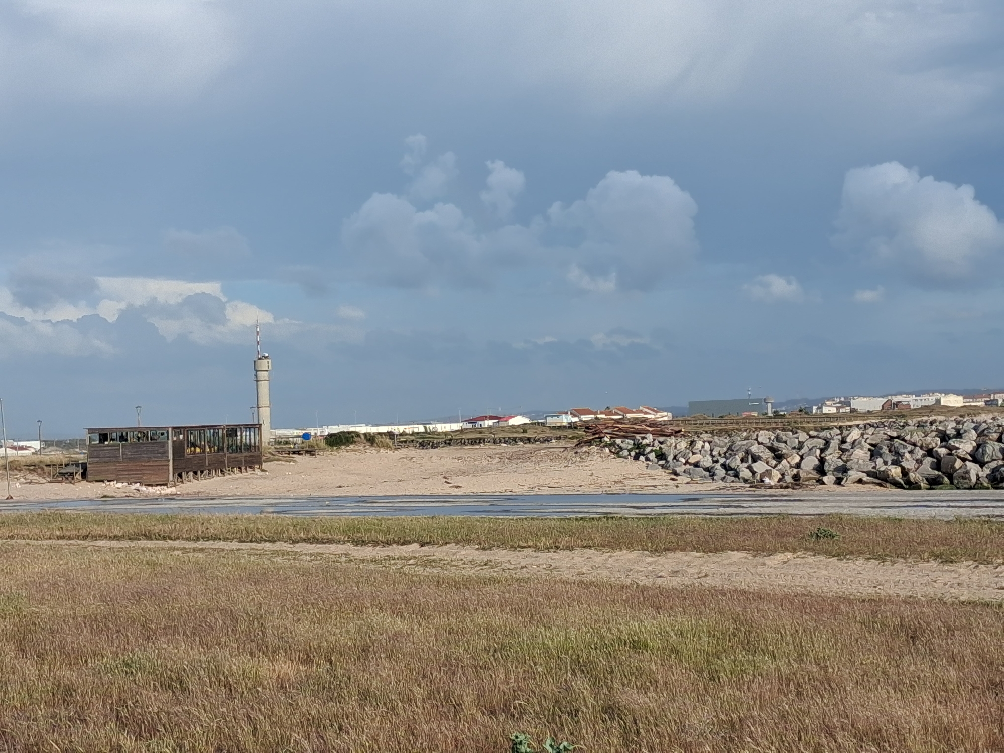 The image depicts a landscape near Figueira da Foz, Portugal, with dry, yellow-brown grass in the foreground. Behind the grass, there's a sandy area leading to a concrete or paved strip. Beyond this, there's a building with a wooden structure and windows, possibly a beach cafe or shelter. A tall, cylindrical tower stands nearby.
In the background, there's a rock barrier, followed by a row of houses and buildings with red roofs. The sky is overcast with patches of blue. No people or animals are visible in the image. The scene appears to be an area where the beach meets a residential or developed area, potentially a coastal zone transitioning from natural landscape to urban infrastructure.
