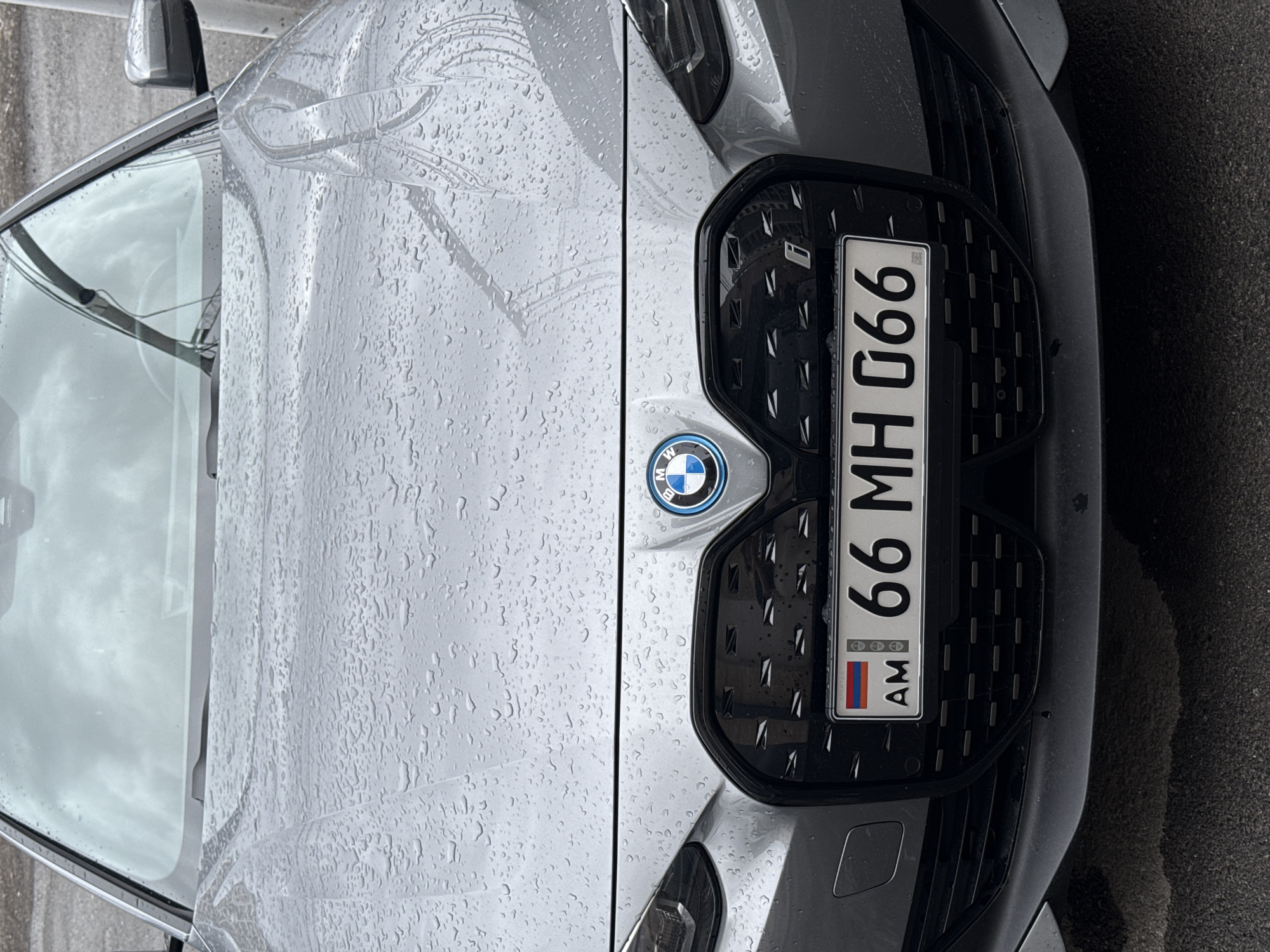 The image displays a close-up, front-facing view of a grey BMW i4 electric vehicle, located in Yerevan, Armenia. The vehicle's exterior, specifically the hood and windshield, is covered in numerous small water droplets, indicating recent precipitation. The BMW emblem, encircled in blue, is centered above a large, black, textured kidney grille that features a subtle "i" badge on its right side. Below the grille, a white rectangular license plate is affixed, displaying the Armenian flag and country code "AM" on the left, followed by the alphanumeric registration "66 MH 066". The windshield reflects an overcast sky. The vehicle is stationary, parked on a dark asphalt surface. No individuals or discernible actions are present in the scene.