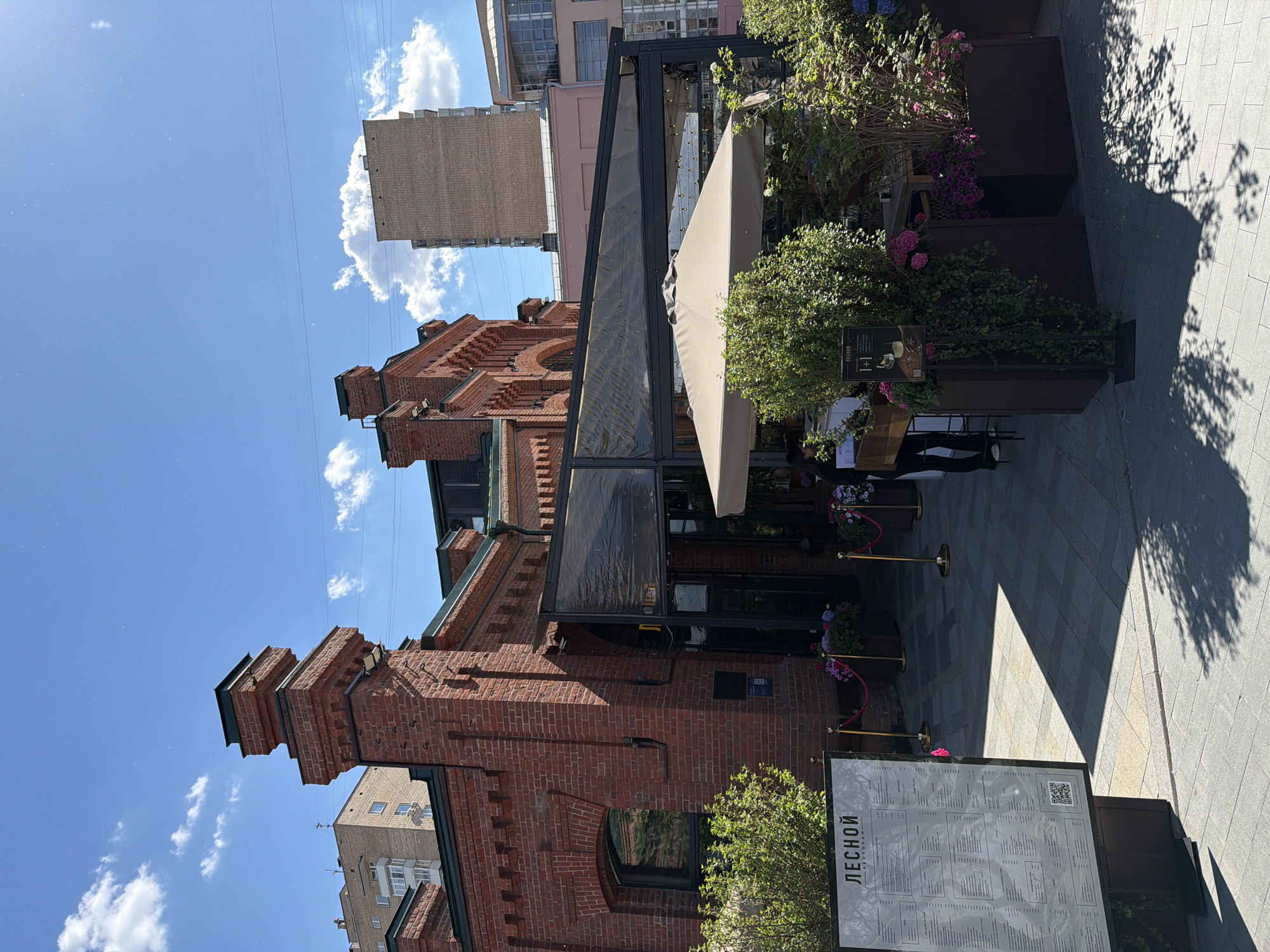 This image captures a bright, sunny outdoor scene in what appears to be a European city, specifically Moscow given the context. The primary subject is a multi-story building constructed from red brick, featuring ornate architectural details like cornices and arched windows. This building, likely an older structure, houses an establishment with an outdoor dining area.
In front of the brick building, an outdoor restaurant space is set up under a dark-framed canopy or awning, partially covered by a large, light-colored umbrella. The area is adorned with numerous planters containing lush green bushes and vibrant pink and purple flowers, creating a pleasant, natural ambiance. Golden stanchions with red ropes define the entrance or a waiting area.
On the paved sidewalk, a large, upright menu board is prominently displayed. The text on the board clearly reads "ЛЕСНОЙ РЕСТОРАН," which translates to "LESNOY RESTAURANT" (Forest Restaurant). A QR code is also visible on the menu.
The time of day is clearly daytime, likely mid-morning or afternoon, characterized by a bright blue sky with scattered white clouds and strong, distinct shadows cast by the trees and buildings onto the pavement, indicating direct sunlight. A few power lines are faintly visible against the sky. In the background, other buildings of varying architectural styles, including a more modern, lighter-colored structure, are visible. Partially visible figures, likely patrons or staff, can be seen near the tables and the menu board, suggesting ongoing activity at the restaurant.