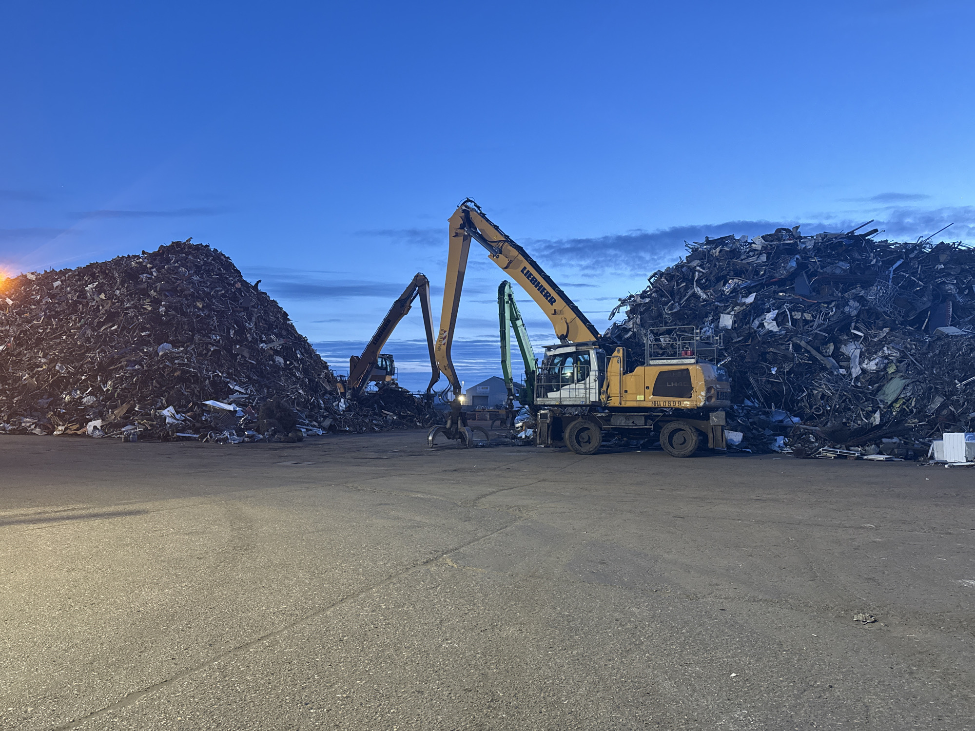The image depicts a scrap metal yard in Gorleston-on-Sea, United Kingdom, at dusk or dawn. Two large piles of scrap metal dominate the scene. Two Liebherr material handler machines are present. One is closer to the foreground and is a yellow machine, and one is farther in the background. The ground is paved. The sky is blue with some clouds.