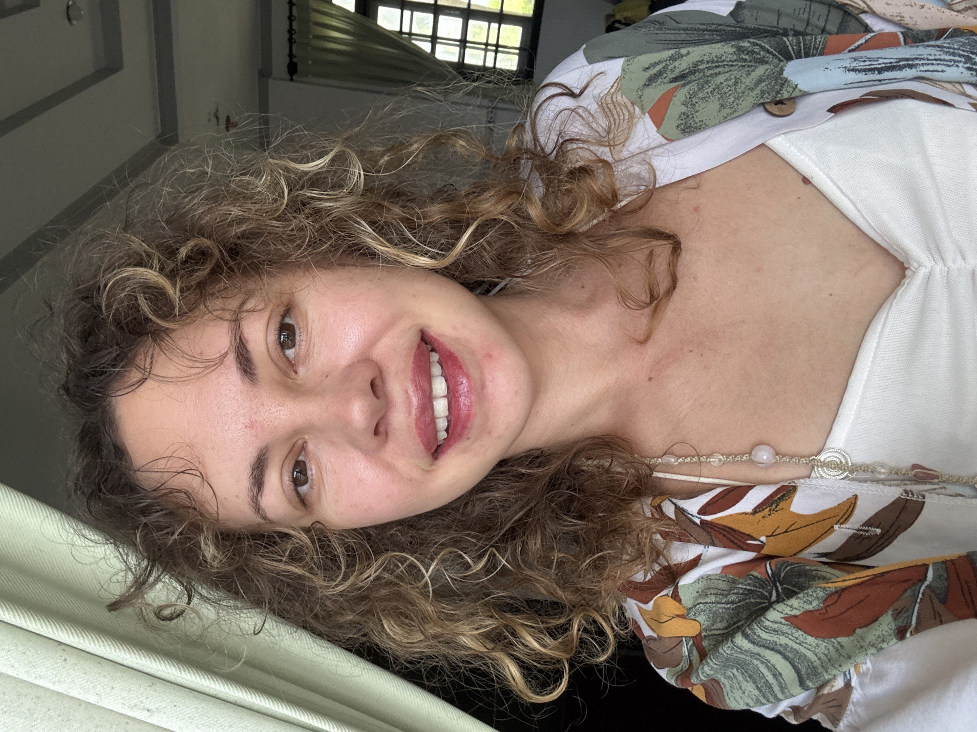 The image shows a close-up, selfie-style shot of a woman with curly, light brown hair. She is wearing a white tank top and a patterned open shirt with large leaves and flowers in shades of green, brown, and orange. A beaded necklace with a coin pendant is visible around her neck. She is smiling with her teeth showing and appears to be indoors, near a window with light-colored curtains. The ceiling appears to be white with recessed lighting. In the background, a window with a grid pattern is visible, showing greenery and a blue sky outside. The overall setting suggests a relaxed, possibly vacation-like atmosphere. Given the context of Mũi Né, Vietnam, the location could be a hotel room, resort, or a similar accommodation with a view of tropical foliage.
