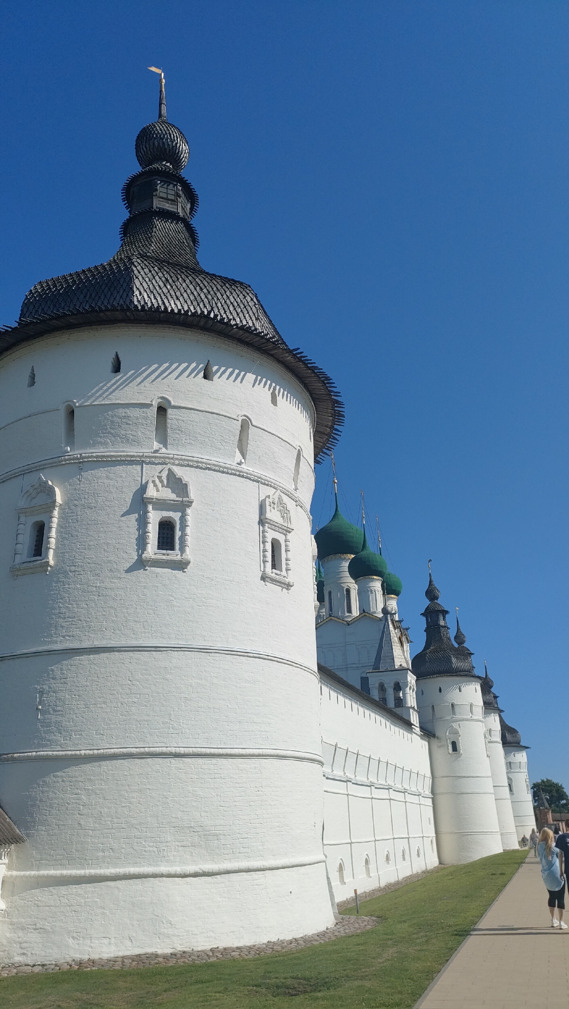 This is a medium-angle, exterior shot of the Rostov Kremlin in Rostov, Russia, taken on a clear, bright day. The main subject is a large, whitewashed brick tower on the left, featuring a dark, shingled conical roof topped with a small, golden weather vane. The tower has several small, rectangular windows with decorative stone surrounds. In the background, a series of whitewashed walls connect to other towers, some topped with dark, pointed roofs, and several green-domed churches are visible. The walls have regularly spaced arched windows. A paved walkway runs along the base of the walls to the right, bordered by a strip of green grass. Two women are walking away from the camera on the walkway. The sky is a deep, clear blue.