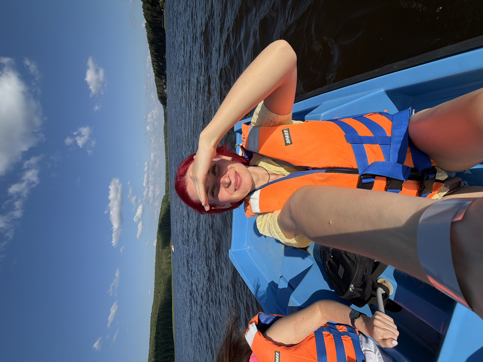 A person with bright red hair, wearing an orange life vest over a yellow shirt, is lying in a blue boat on a body of water. They are shielding their eyes from the sun with their hand and smiling at the camera. The boat appears to be moving, as indicated by the ripples on the water.
In the background, a dense, green forest lines the shore of the lake. The sky is a clear, bright blue with a few fluffy white clouds. The sun is shining, suggesting it is daytime and the weather is fair.
Another person's arm, also wearing an orange life vest, is visible in the foreground, holding onto a gray handle. A black bag is positioned behind them in the boat. The overall scene suggests a leisurely activity, likely a boat trip or watersport on a sunny day.
The text "SPASS" is visible on the orange life vest. The location is identified as Chaykovsky, Russia.