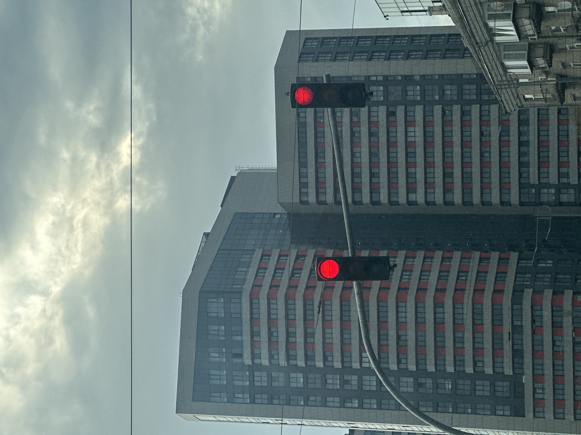 The image shows a low-angle view of two modern buildings in Kyiv, Ukraine. The buildings are tall and have a repetitive pattern of windows and red vertical stripes. A traffic light with two red lights is visible in the foreground. The sky is overcast and grey. Overhead power lines are visible.